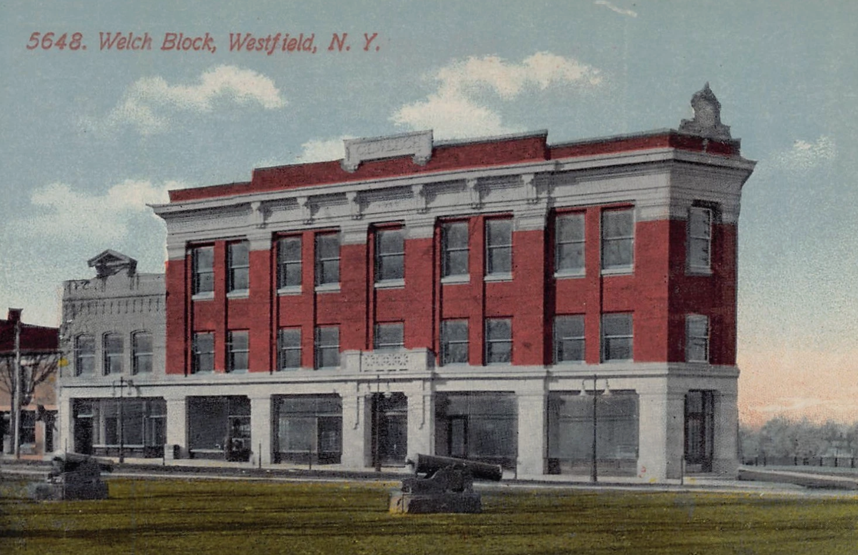 A historic red brick building with white trim, labeled 5648 Welch Block, Westfield, N. Y., set against a blue sky with clouds.