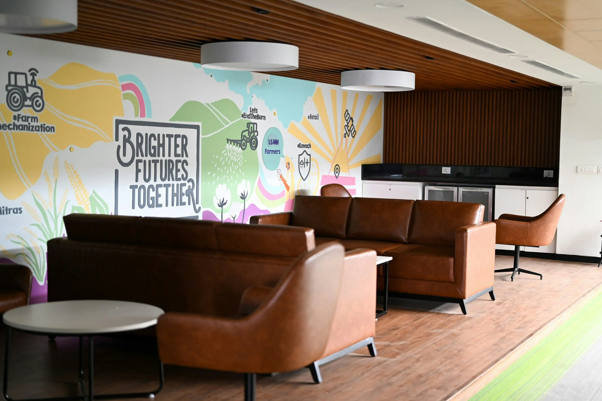 Interior view of a modern lounge area with brown leather chairs, white circular tables, and a colorful mural on the wall featuring illustrations of farming, technology, and renewable energy symbols with the phrase 'Brighter Futures Together.' The ceiling has wooden slats and two white hanging pendant lights.