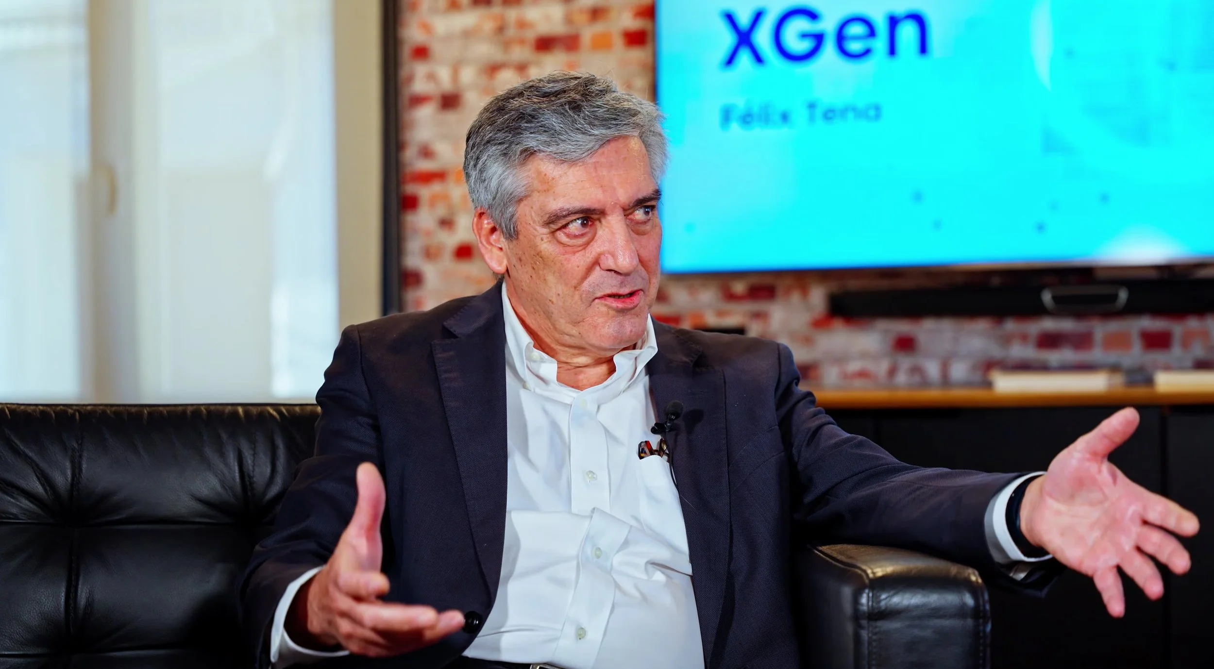 Hombre con cabello gris en una conferencia, usando saco y camisa blanca, gesticulando con las manos. En la pantalla detrás, el texto 'XGen' y 'Folix Tano'.