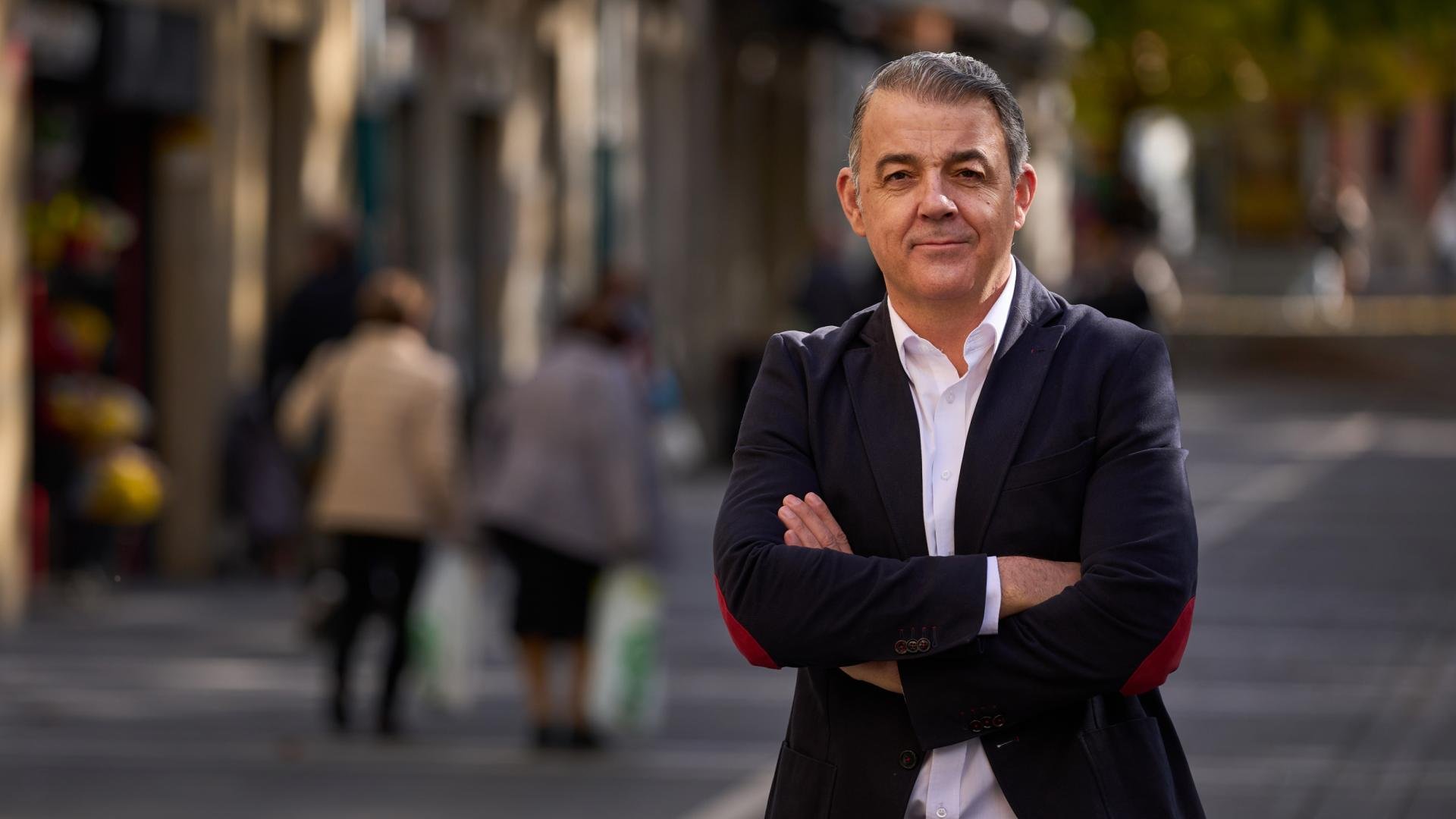 Hombre con cabello gris y ropa formal posando con brazos cruzados en la calle, fondo difuso con otras personas y árboles.