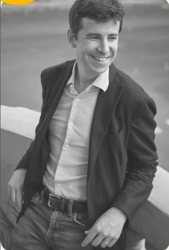 Joven sonriendo, usando blazer y camiseta, posando cerca de una baranda al aire libre.