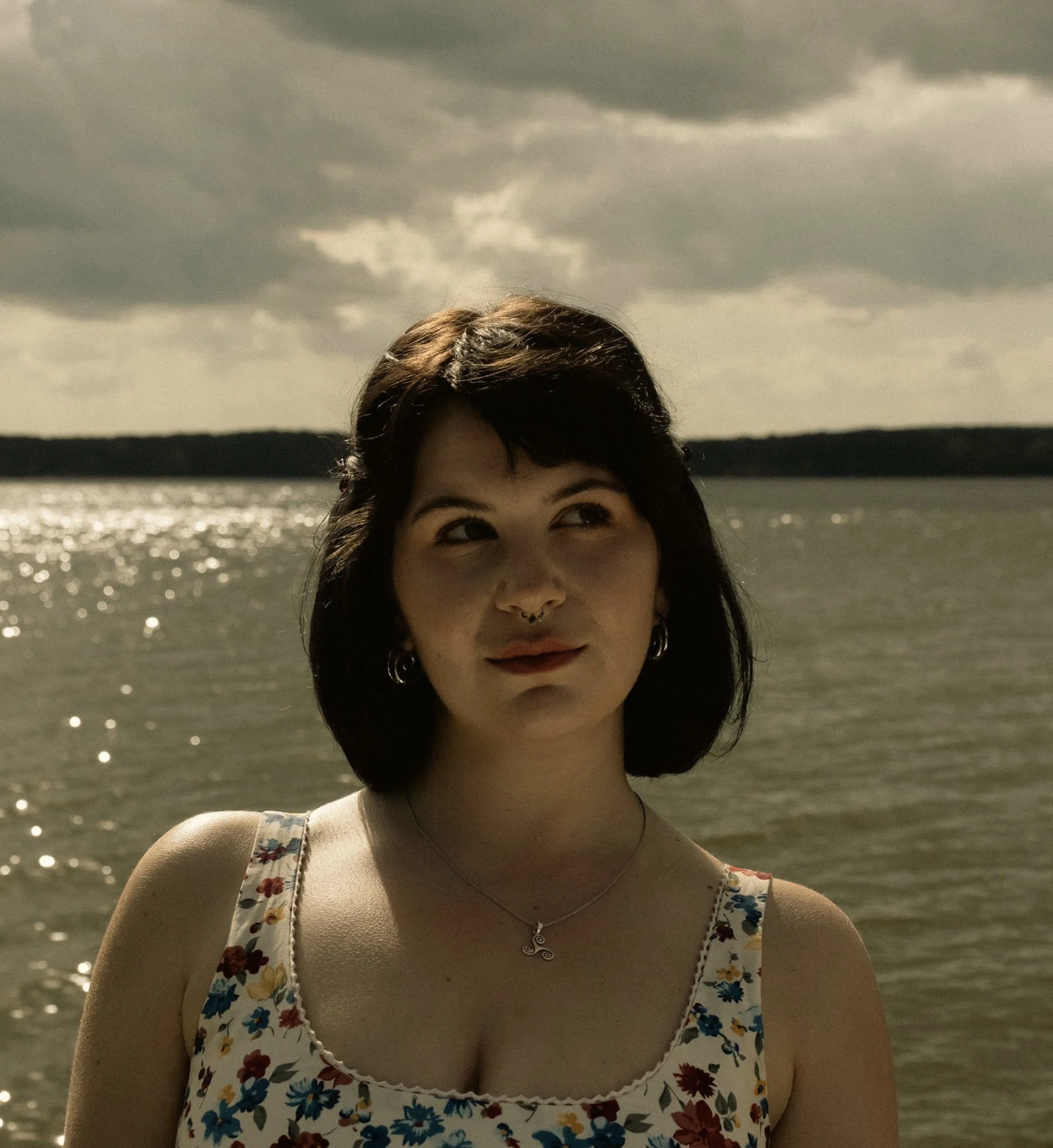 A woman with dark hair is near a body of water, looking slightly upwards, during cloudy weather.