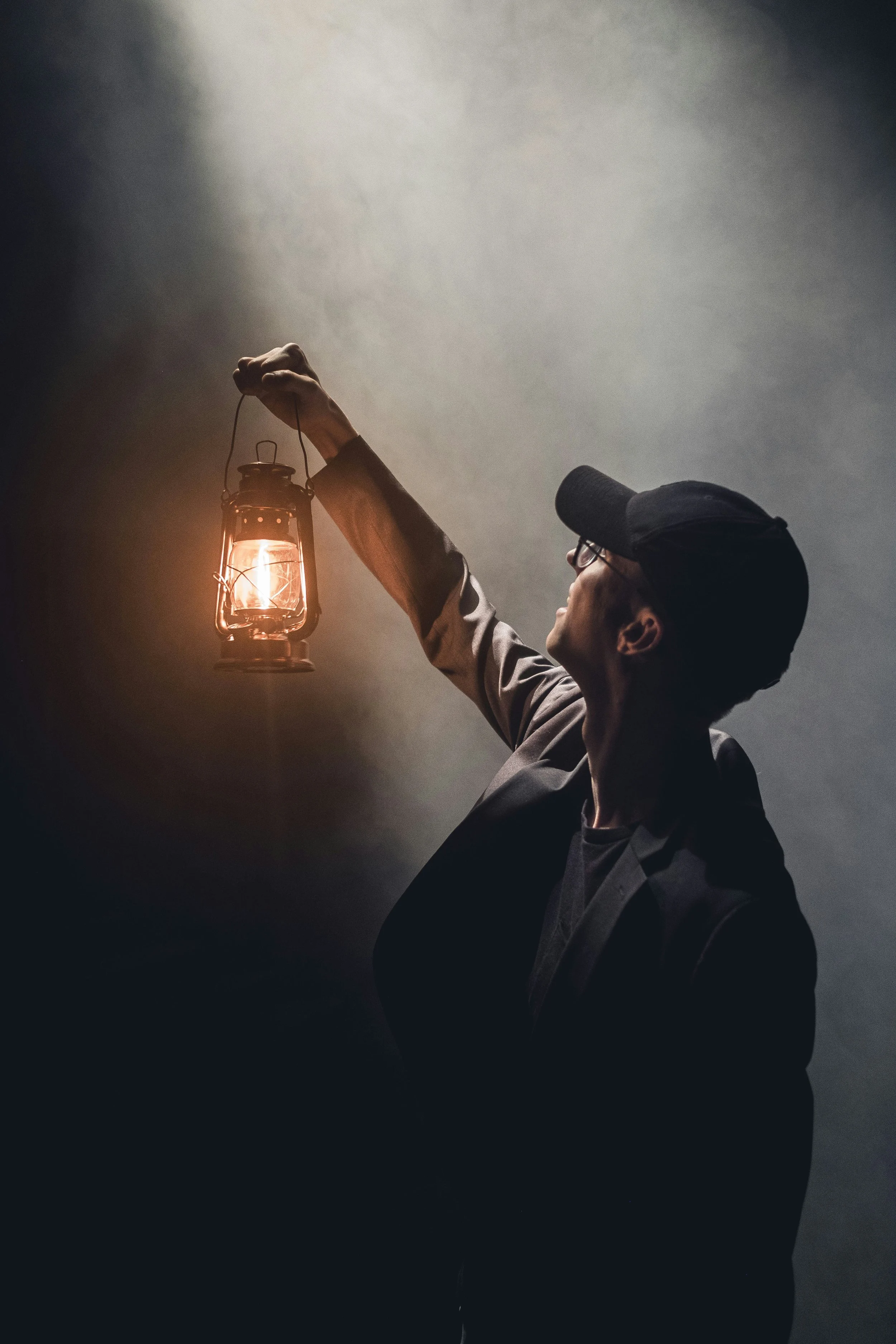 A person wearing glasses and a black cap holds up a lit lantern in a dark, smoky environment.