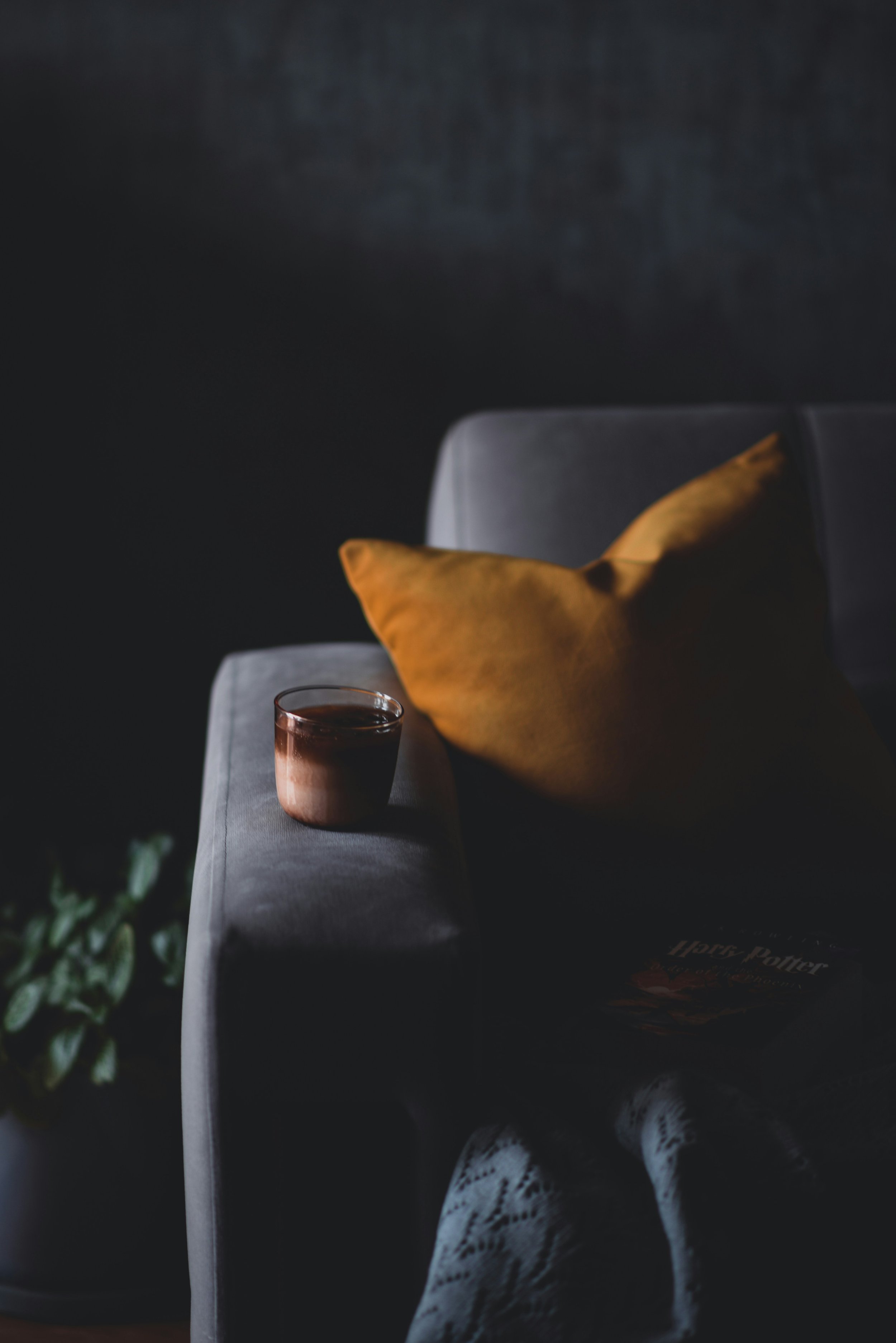 Close-up of a grey sofa with a mustard yellow pillow and a glass of dark beverage on a grey armrest, dark background, person wearing white lace shoes at the bottom.
