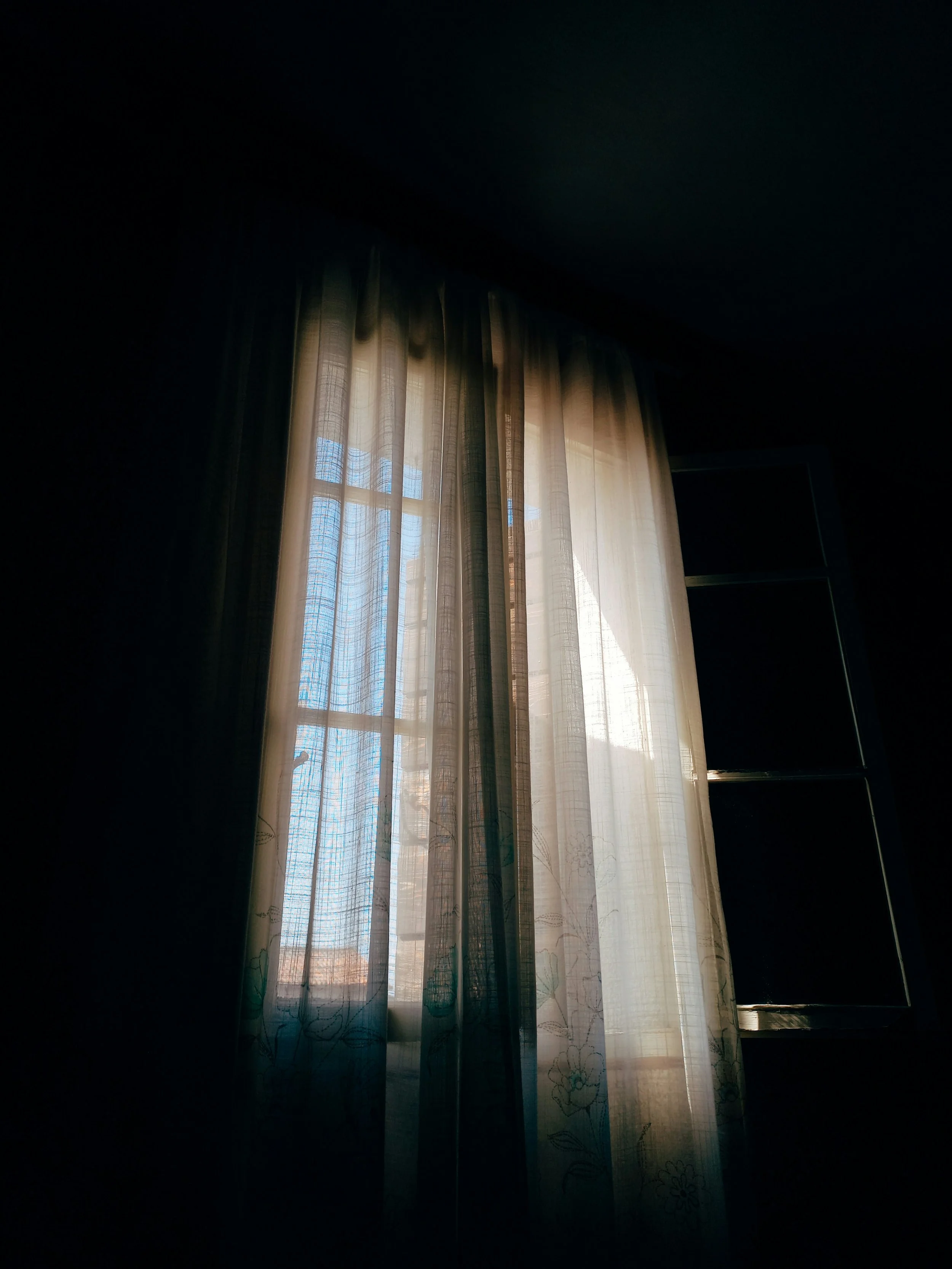Sunlight filtering through sheer curtains in a dark room, with a view of a blue sky and trees outside.