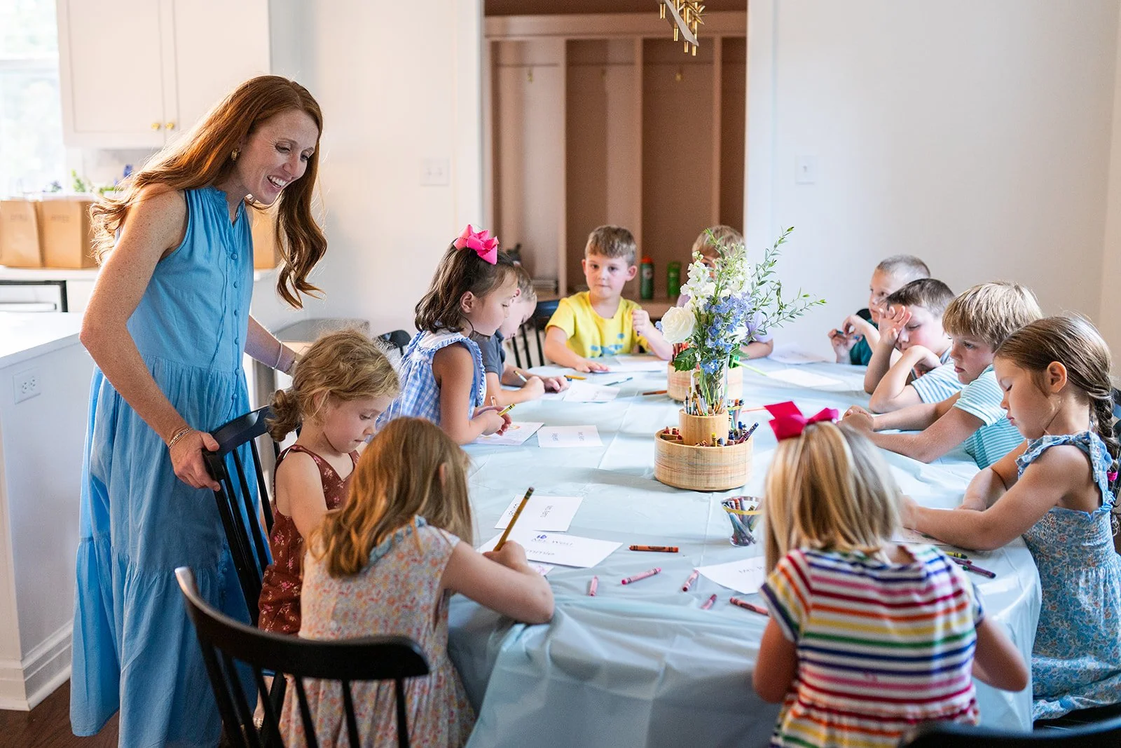 A young woman in a blue dress stands at the head of a long table with children seated on both sides, involved in drawing and coloring activities. The setting appears to be a lively children’s party or classroom, with a centerpiece of flowers in the middle of the table.