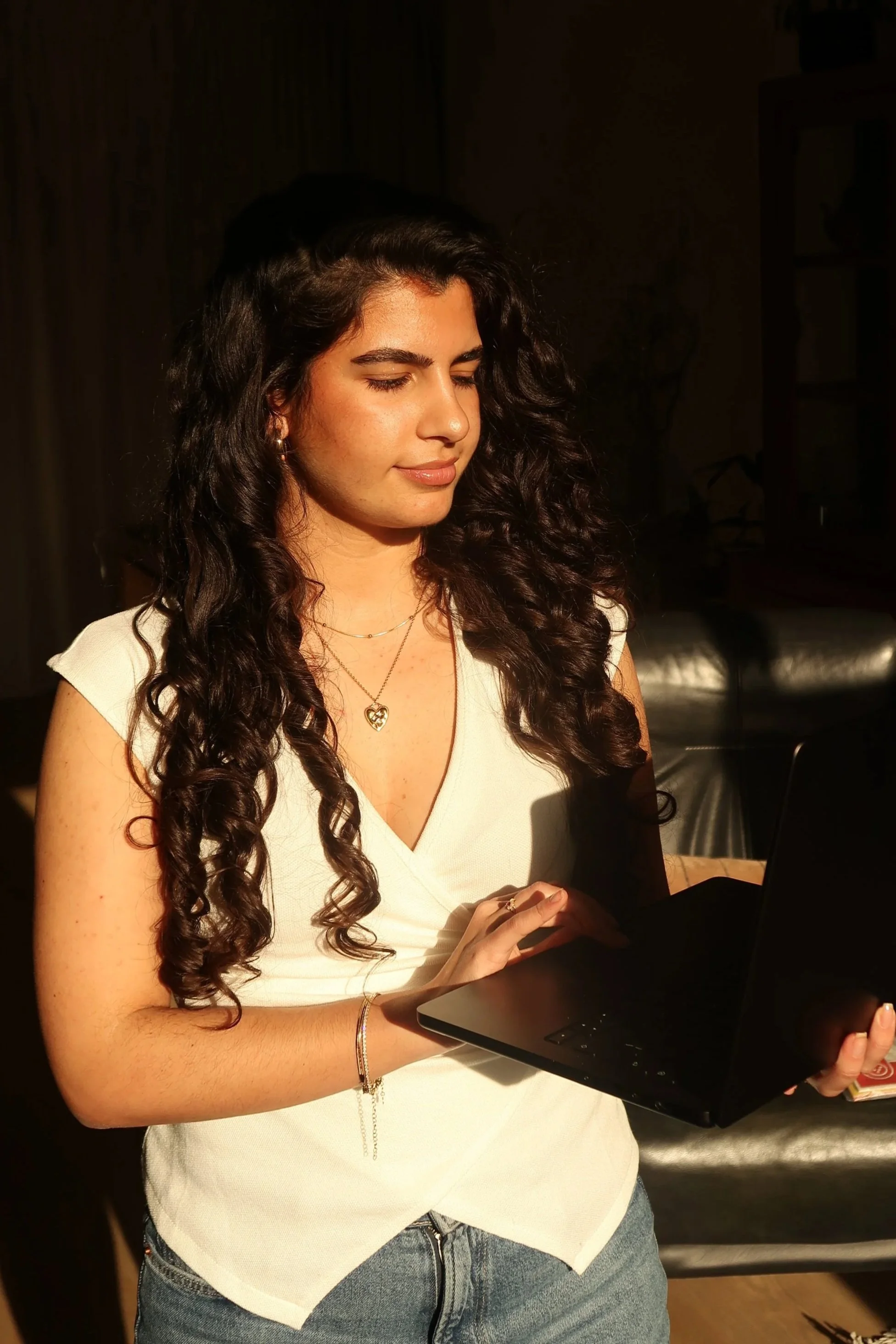 Een jonge vrouw met lang, gekruld bruin haar, een lichte top en een ketting met een hartvormige hanger, die op een laptop typt of iets erop vasthoudt, in een binnenruimte met dimme verlichting.