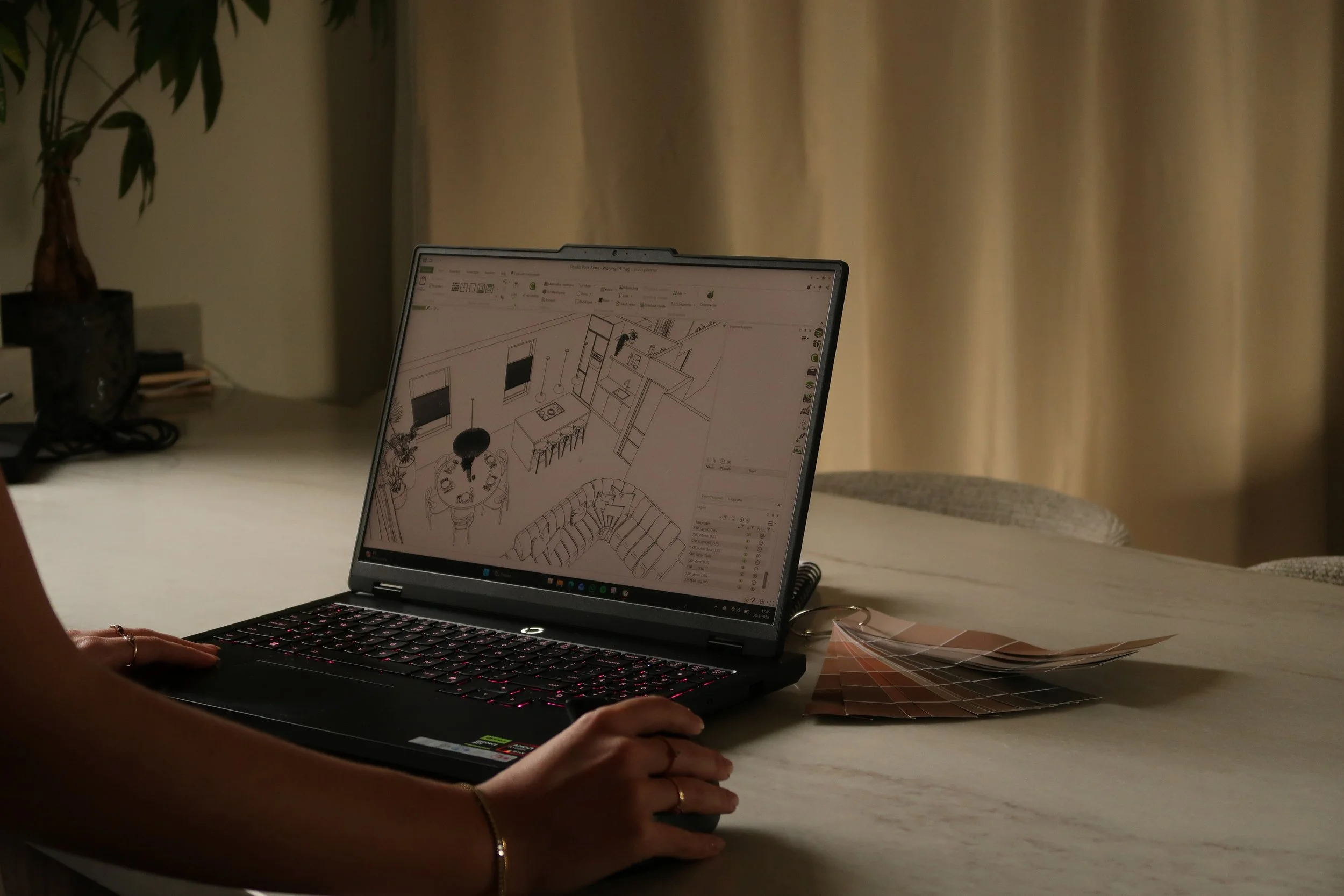 Een persoon werkt op een laptop met een schetsontwerp van een interieur. Op de tafel liggen kleurstalen, en op de achtergrond hangt een beige gordijn.
