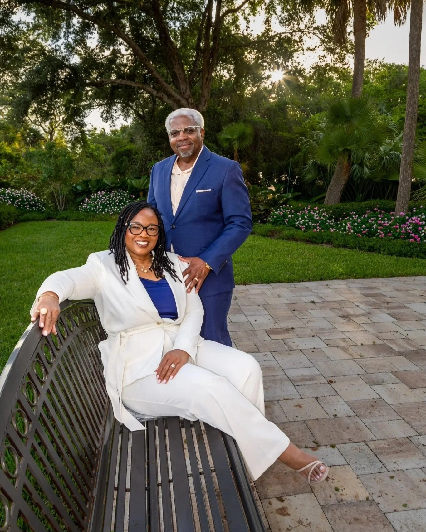 A woman in a white suit sitting on a bench and a man in a blue suit standing beside her outdoors with trees and bushes in the background, at sunset.