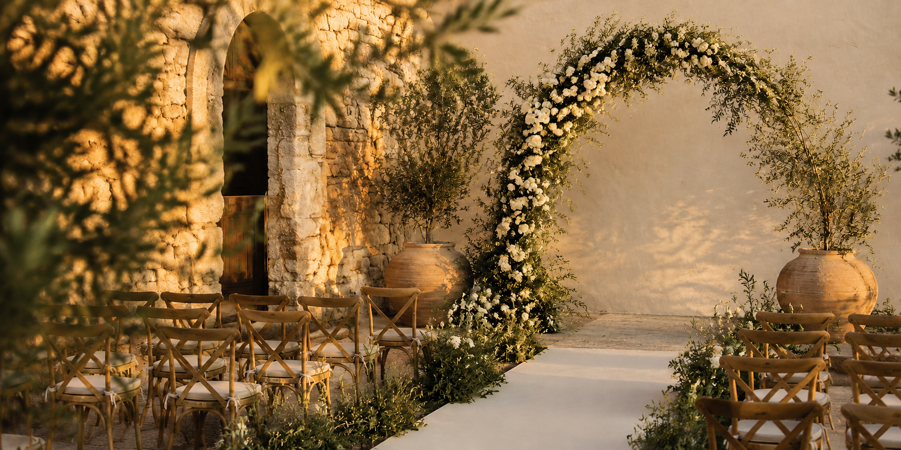 An outdoor wedding ceremony setup with wooden chairs arranged in rows on a white aisle runner, flanked by potted plants and a floral arch made of white flowers and greenery. The scene is illuminated by warm sunlight, with a stone wall and large vases in the background.