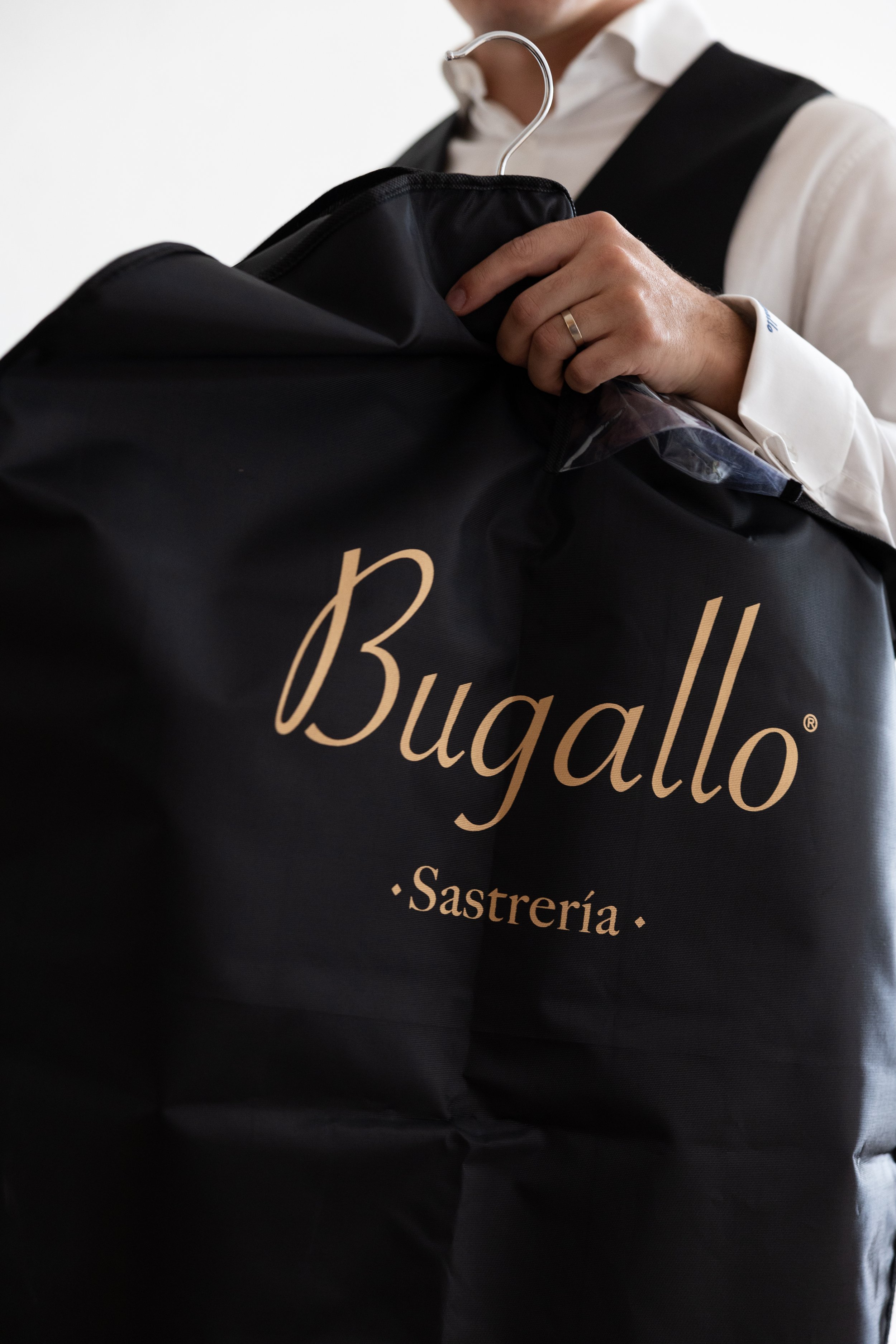 Person in a white shirt and black vest holding a black bag with gold lettering that reads 'Bugallo Sastrería'.