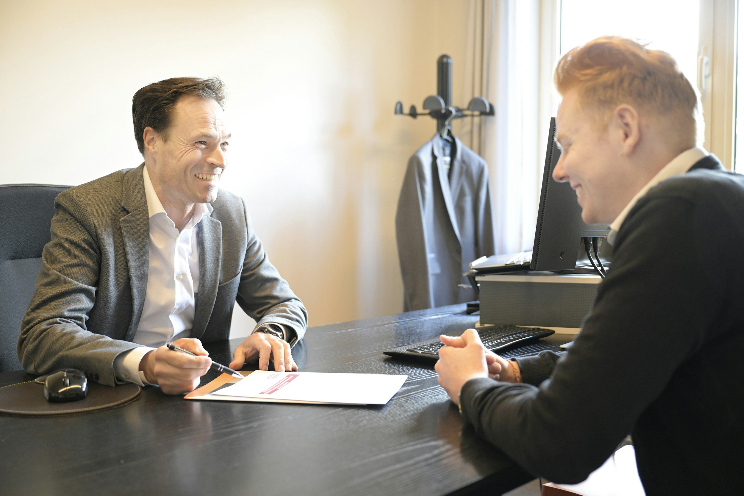 Two men in business suits sitting across from each other at a table in a bright office. One man, holding a pen, is smiling and appears to be in a conversation with the other, who is smiling with his hands clasped on the table. A computer monitor and office supplies are on the desk.