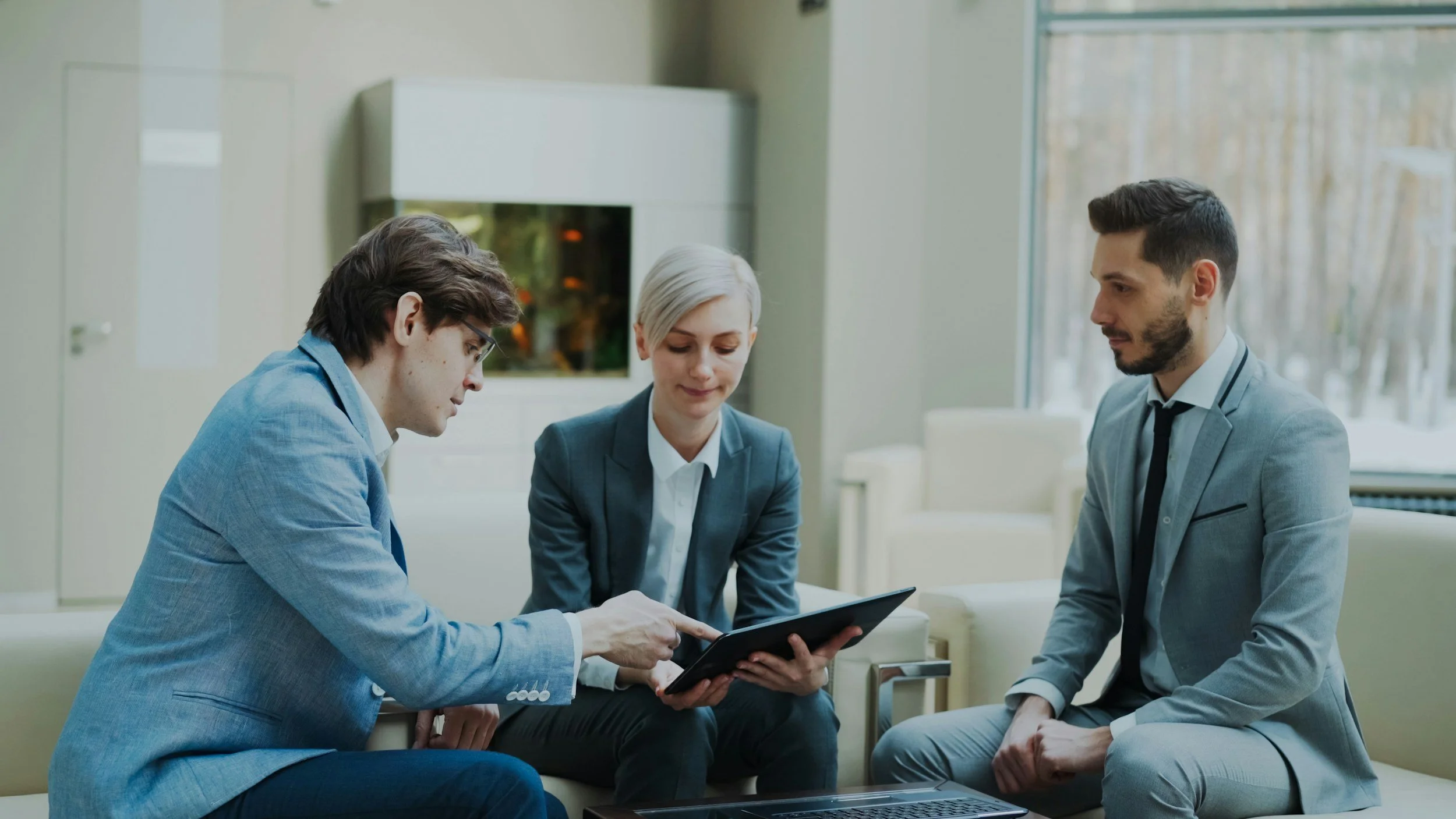 Four professionals, two men and two women, in business attire, engaged in a discussion, with one man showing a tablet to the group in a modern office setting.