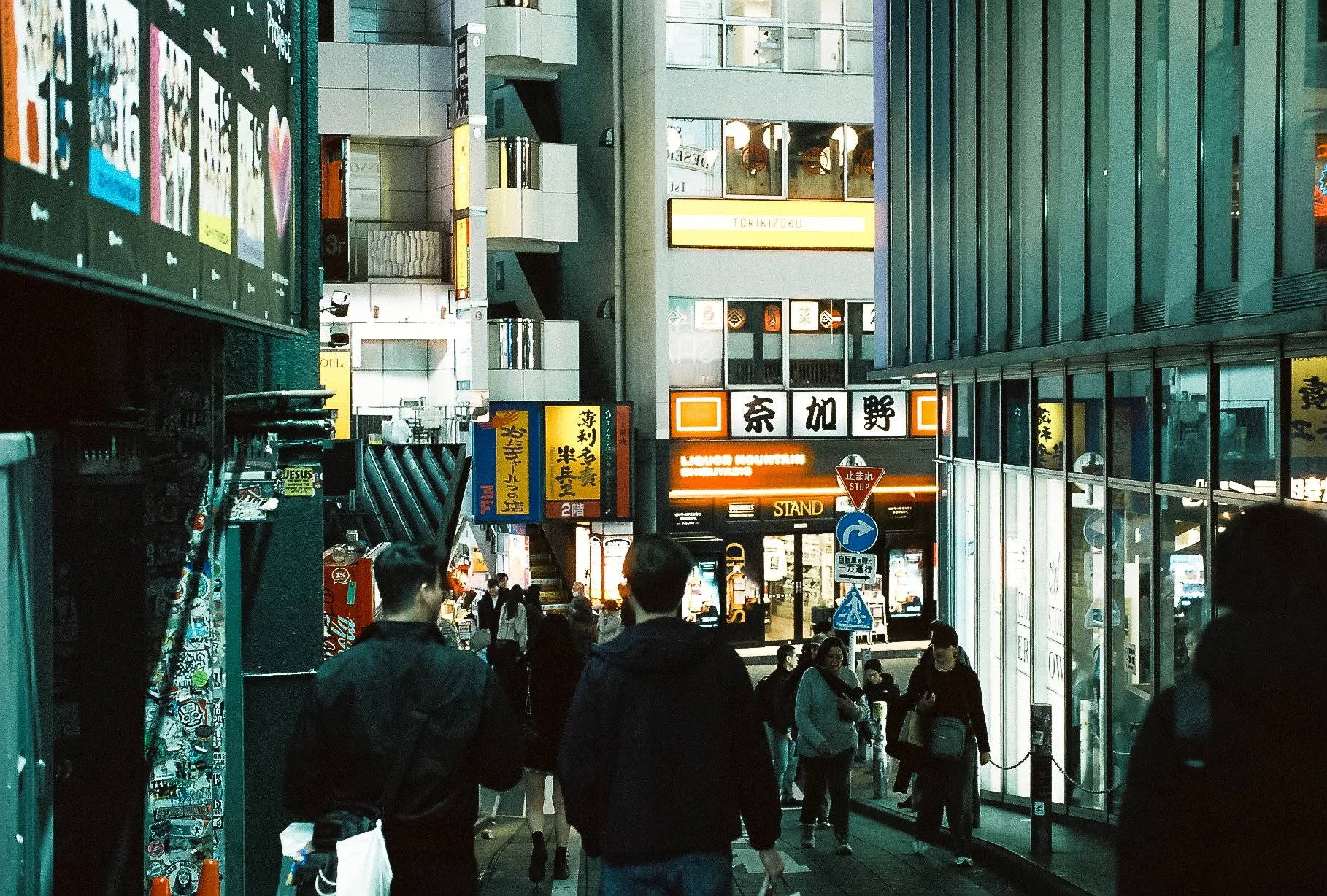 Urban night photography looking down a bustling pedestrian sidewalk lined with glowing storefronts in Japan.