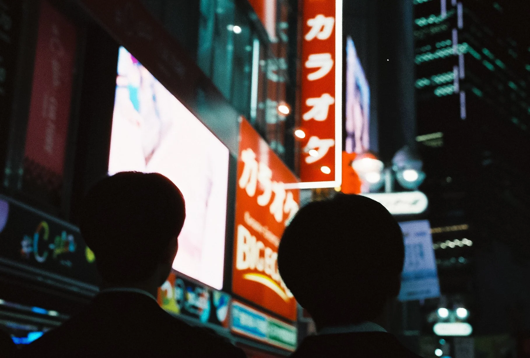 Silhouettes of people walking past a bright red illuminated Japanese karaoke sign at night.