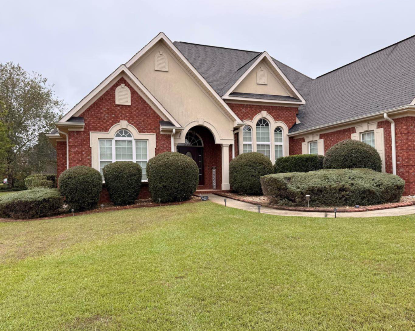 A large two-story house with a brick exterior, multiple arched windows, and a black front door, surrounded by well-manicured bushes and a green lawn.