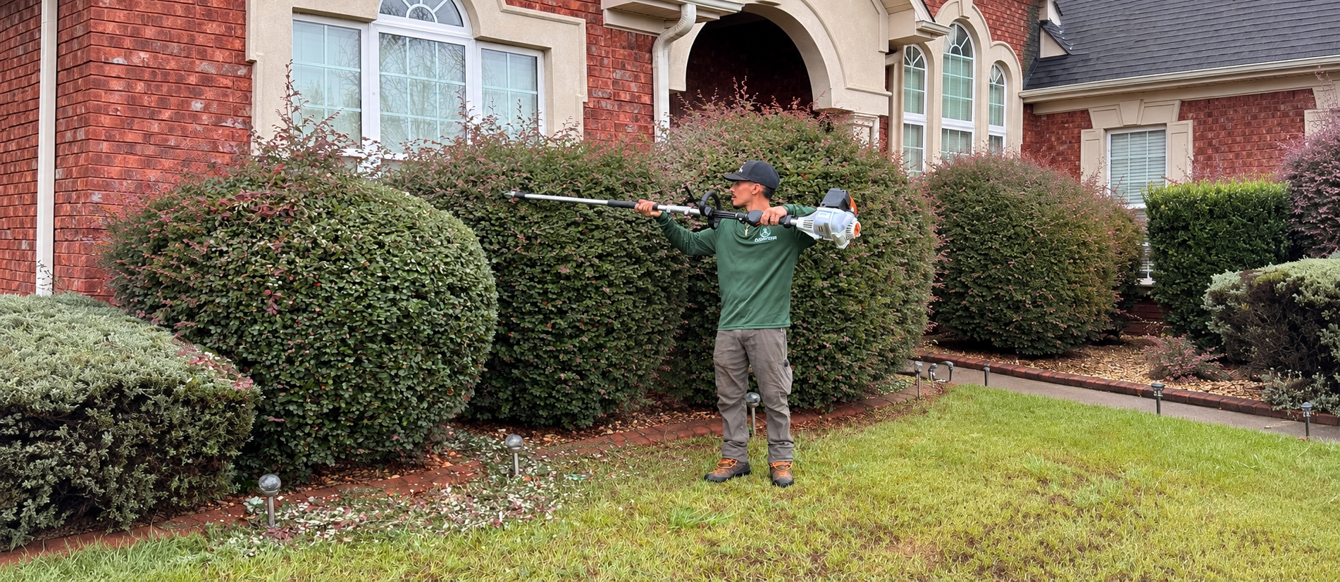 A man using a leaf blower to clean up flower petals in front of a brick house with large bushes and windows.