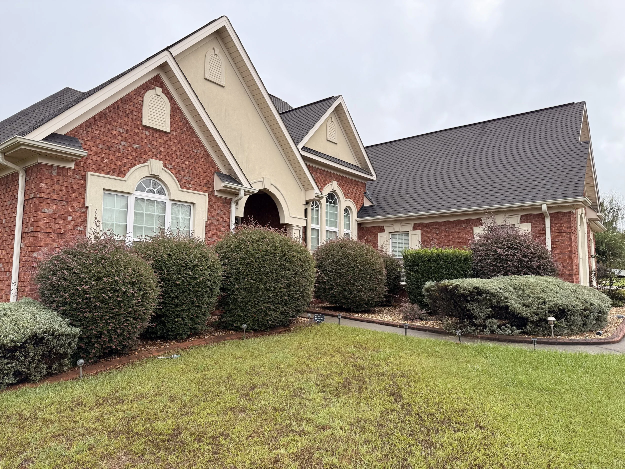 A large, upscale house with red brick and beige siding, multiple gabled roofs, and white window shutters. The front yard features manicured bushes, small landscape lights, and a lush green lawn under an overcast sky.