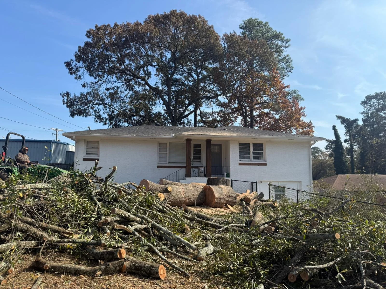 A white house with a porch, surrounded by large fallen tree logs and debris in the foreground, with trees and a blue sky in the background.