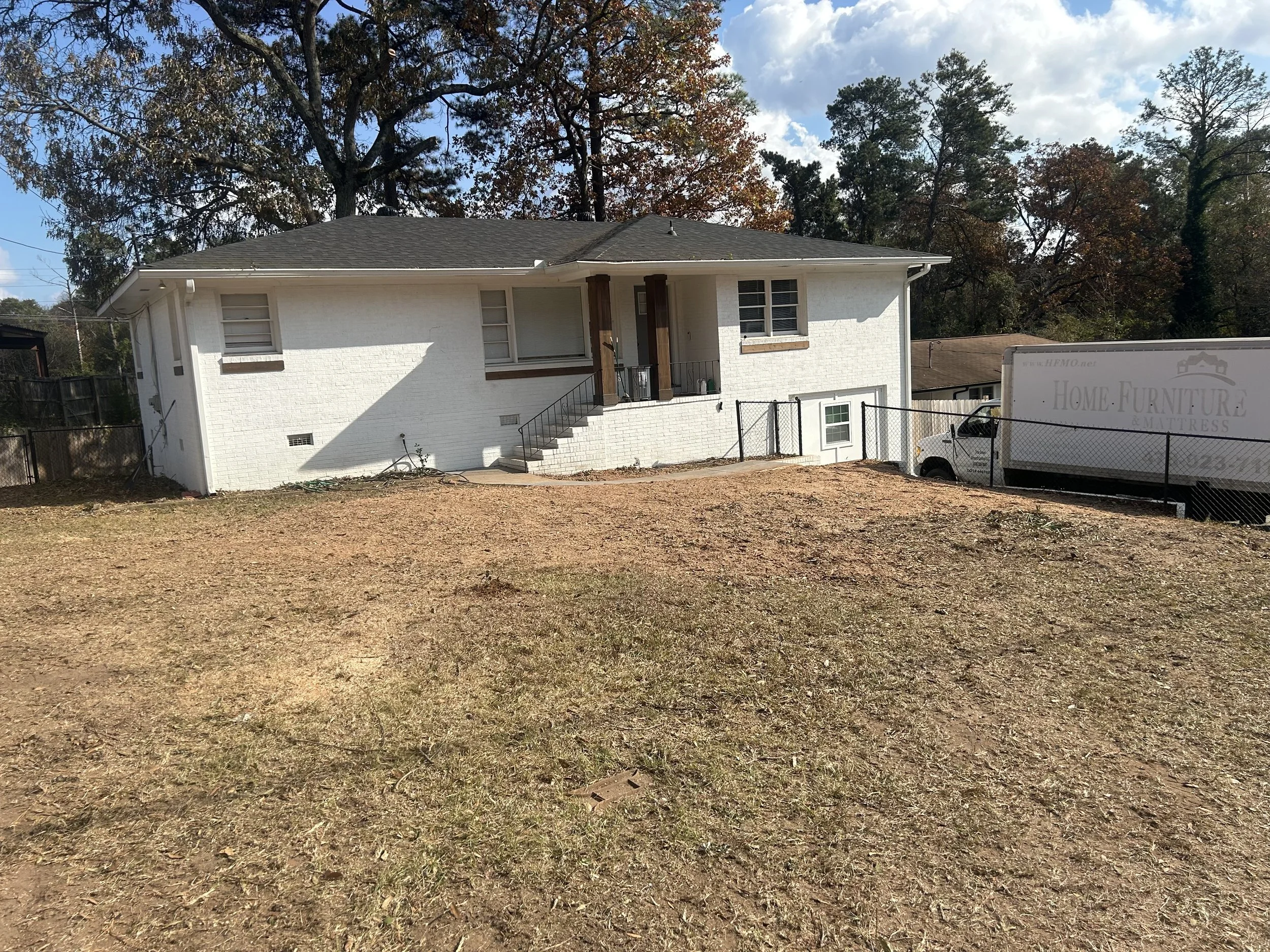 The photo features a two-story house with white brick exterior walls, a black roof, and a small front porch with steps. There are trees behind the house with autumn-colored leaves, and a truck with logo parked on the right side of the property.