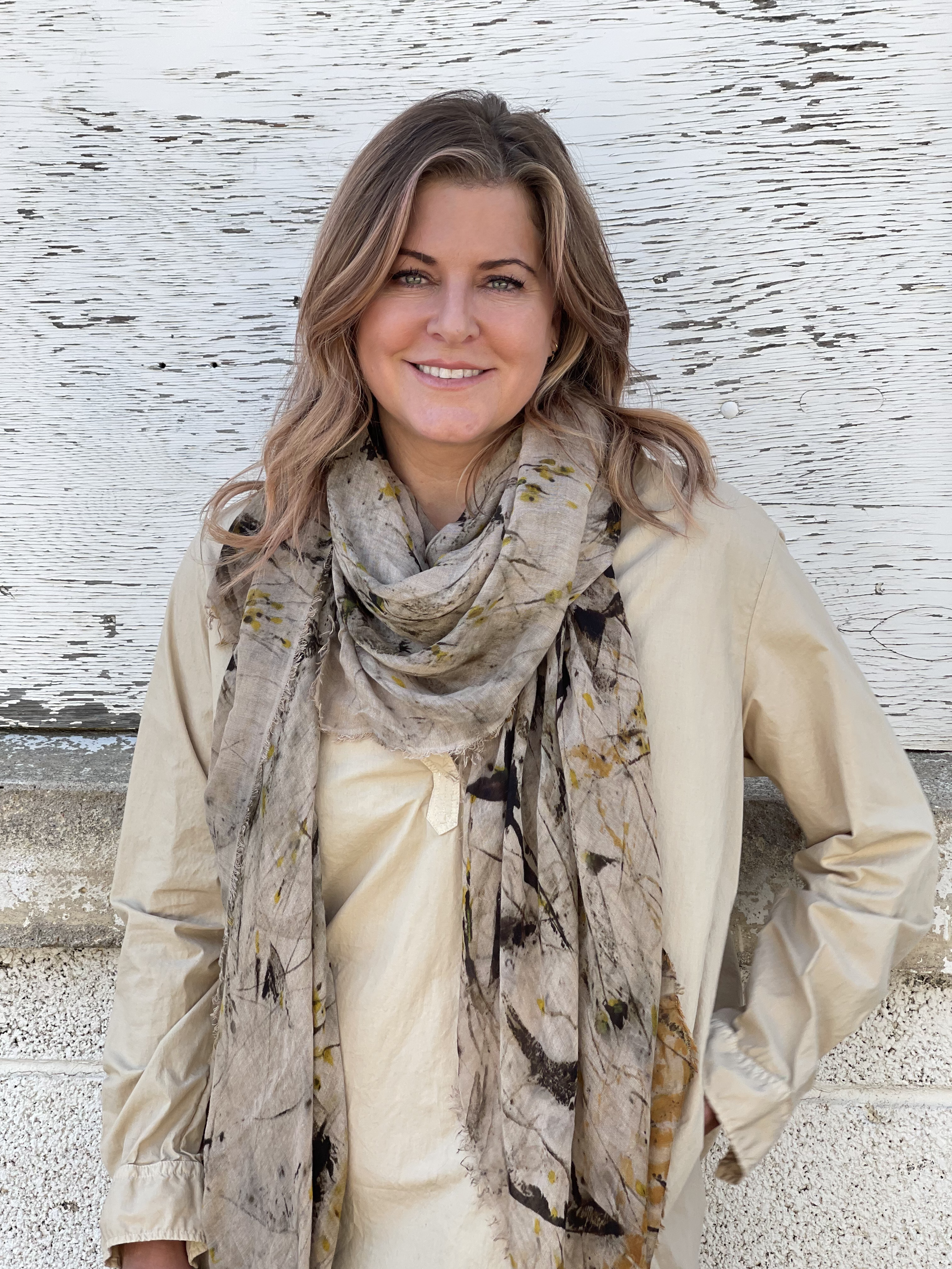 Portrait of a woman with wavy light brown hair, light skin, and green eyes, smiling, wearing a beige jacket and a floral scarf, standing outdoors in front of a white wooden wall.