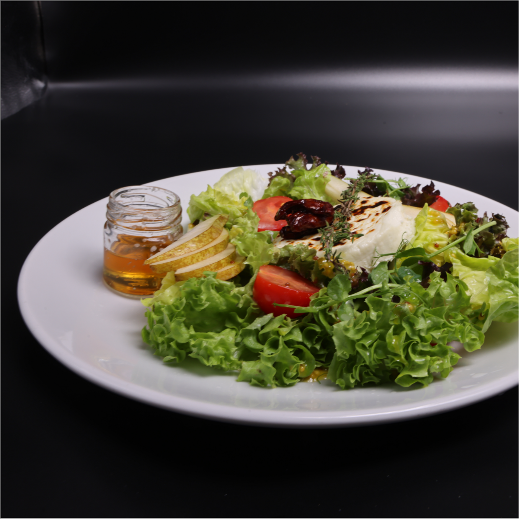 A plate of fresh green salad with tomatoes, lettuce, cheese, sun-dried tomatoes, and herbs, served with a small jar of honey or dressing, on a white plate against a black background.