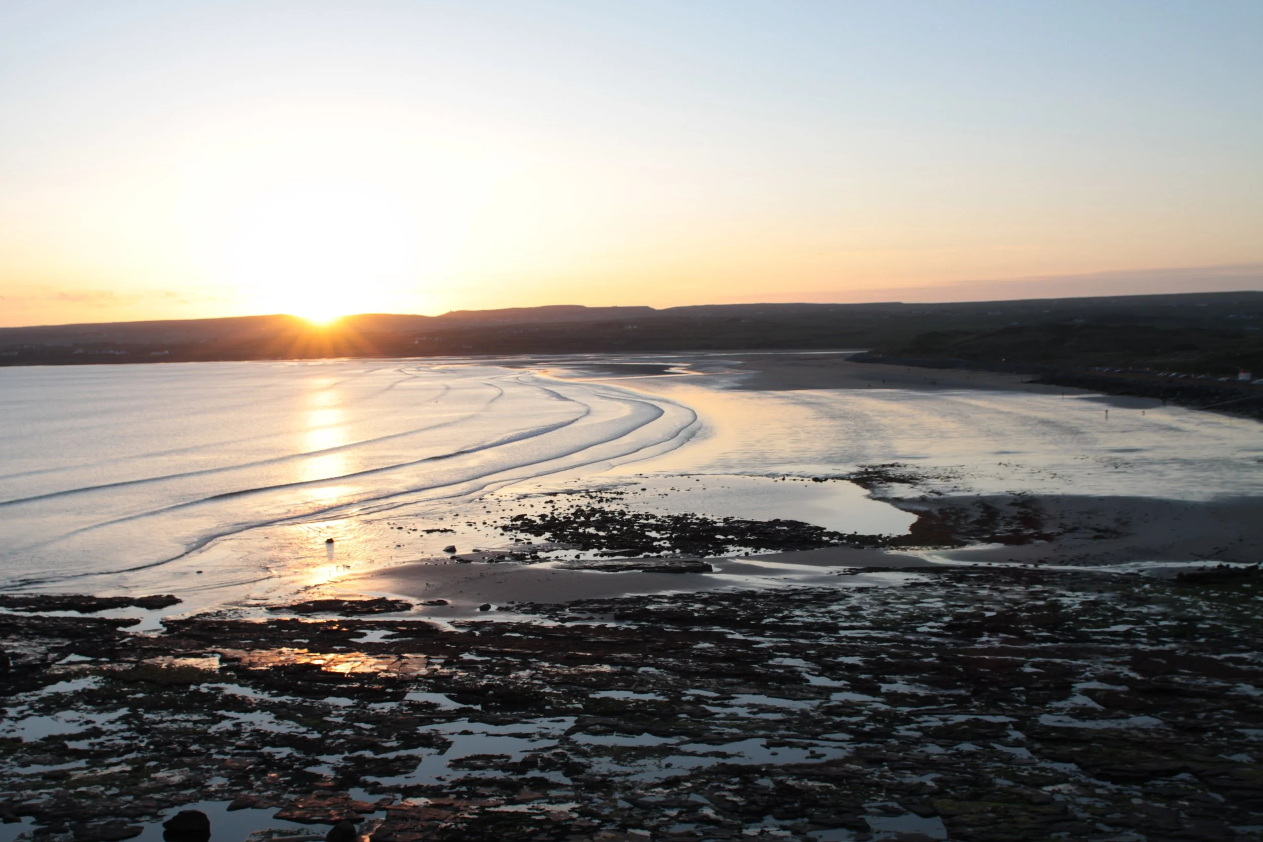 Sunset over Lahinch Beach with small waves and rocky shoreline in foreground.