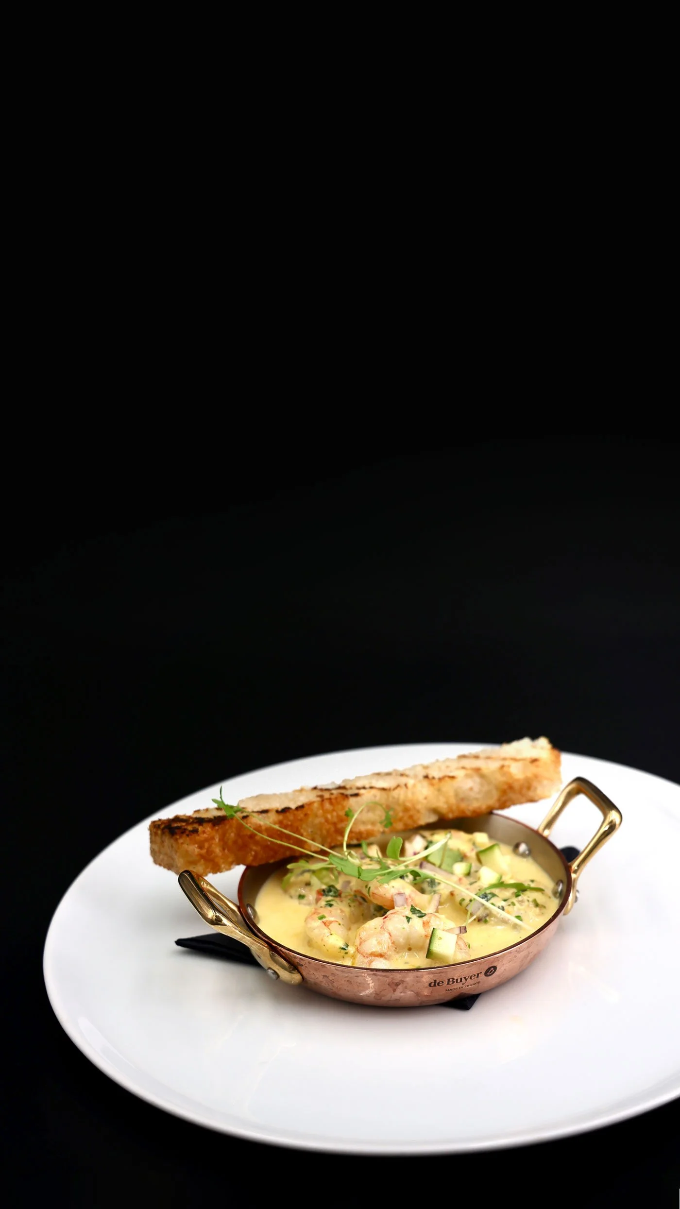 Shrimp and vegetable chowder in a copper bowl garnished with herbs, with a slice of toasted bread on a white plate against a black background.