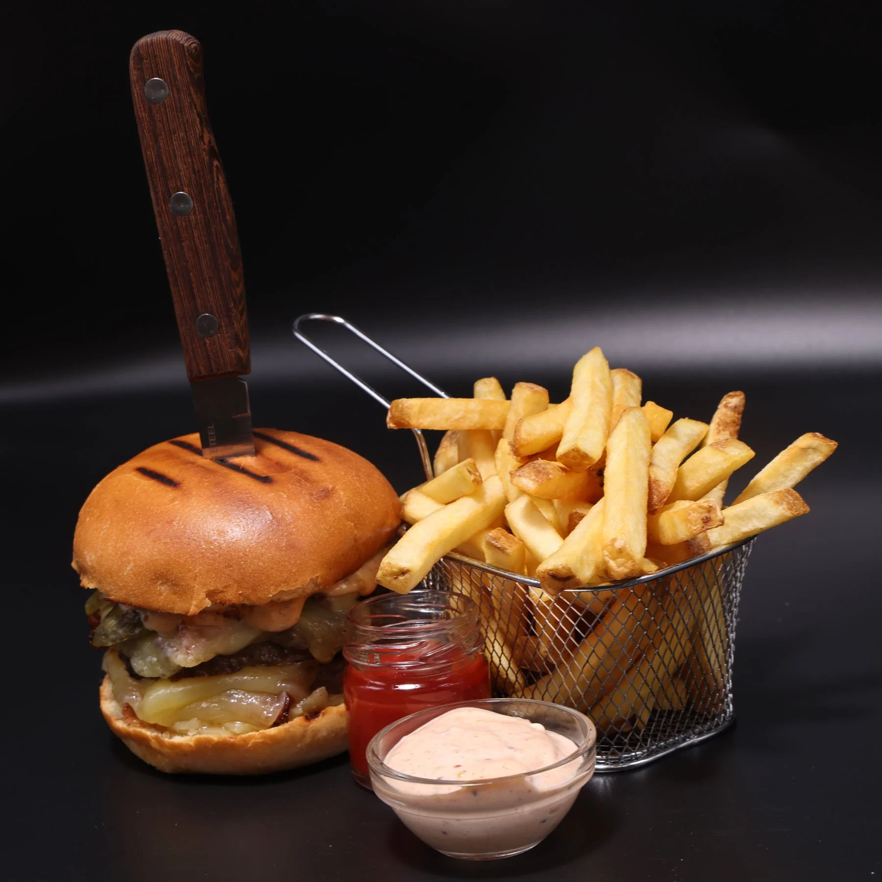 A cheeseburger with a bun, melted cheese, beef patty, bacon, and pickles, accompanied by a basket of French fries, ketchup, and mayonnaise, set against a black background.