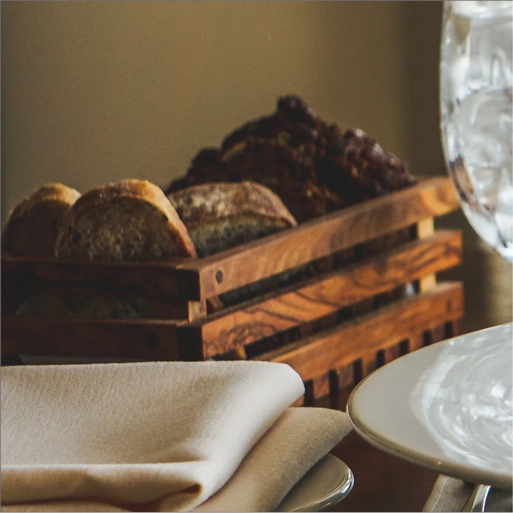 A wooden tray filled with various freshly baked bread rolls and slices, placed on a table with a white napkin, glassware, and a decorative glass pitcher nearby.