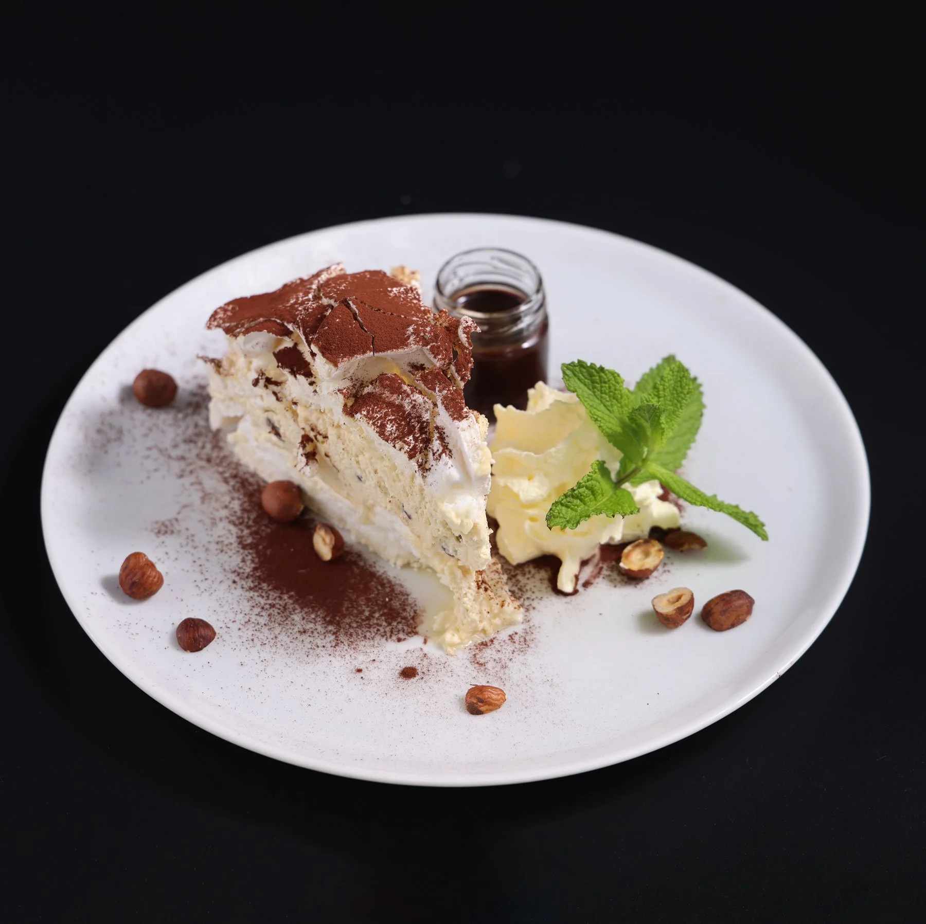 Slice of tiramisu with cocoa powder, hazelnuts, a small jar of sauce, whipped cream, and mint on a white plate against a black background.