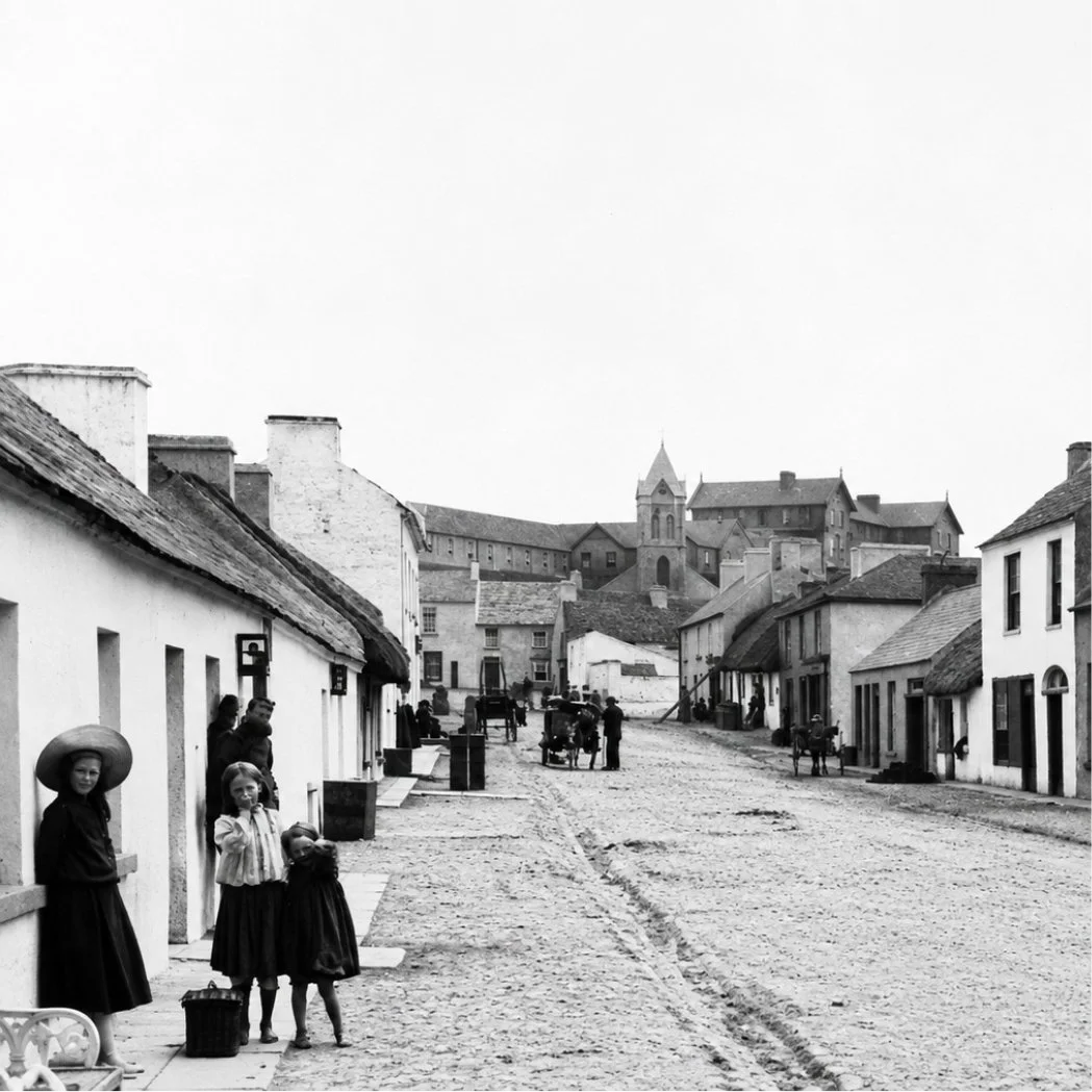 A black and white photo Main Street Lahinch with dirt road and old buildings. Children and women with baskets are standing outside houses, some conversing. In the background, there are more buildings and a church with a steeple.