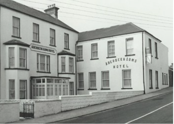 Black and white photo of the exterior of the Abedeen Arms Hotel, a multi-story building with several windows and a small front yard enclosed by a low wall, located on a sloped street in Lahinch, Co. Clare