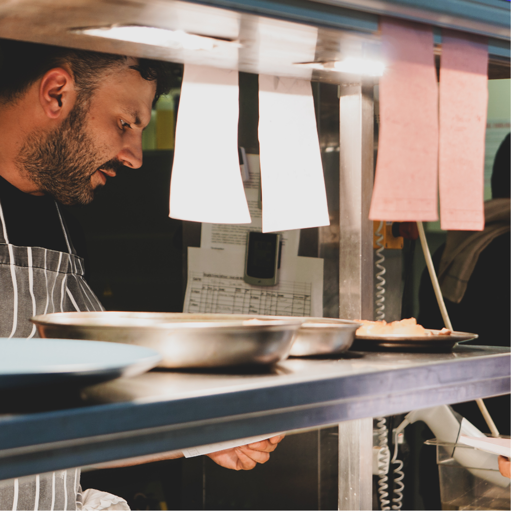 A man in a striped apron working in a kitchen or food service setting, seen through a gap in a stainless steel counter and pink and white papers hanging above him.