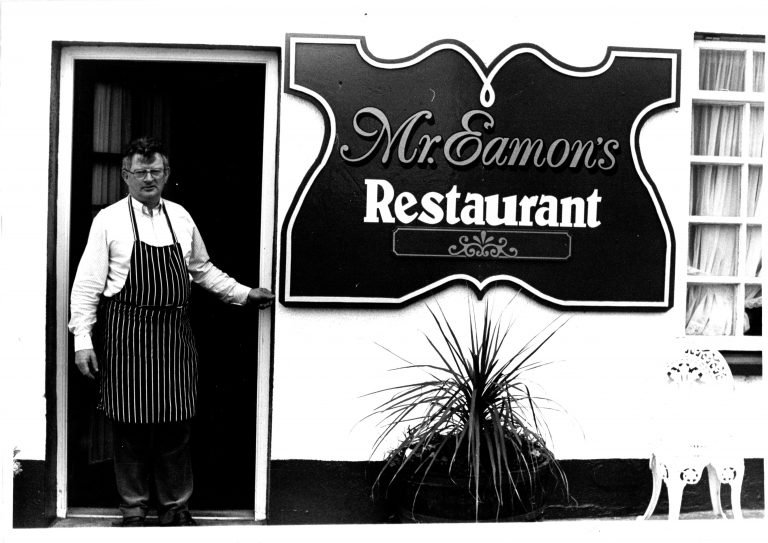 Mr Eamon in a striped apron standing at the entrance of Mr. Eamon's Restaurant, with a sign above him displaying the restaurant's name.