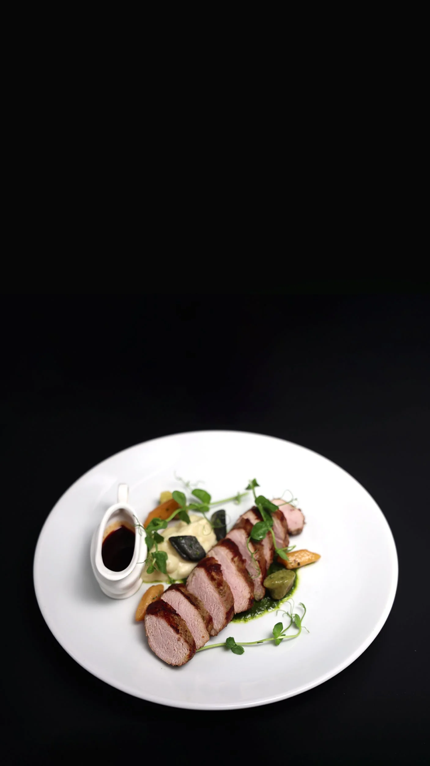 A white plate of sliced cooked meat, garnished with herbs, alongside a small jug of dark sauce, mashed potatoes with green garnish, and vegetables, presented on a black background.