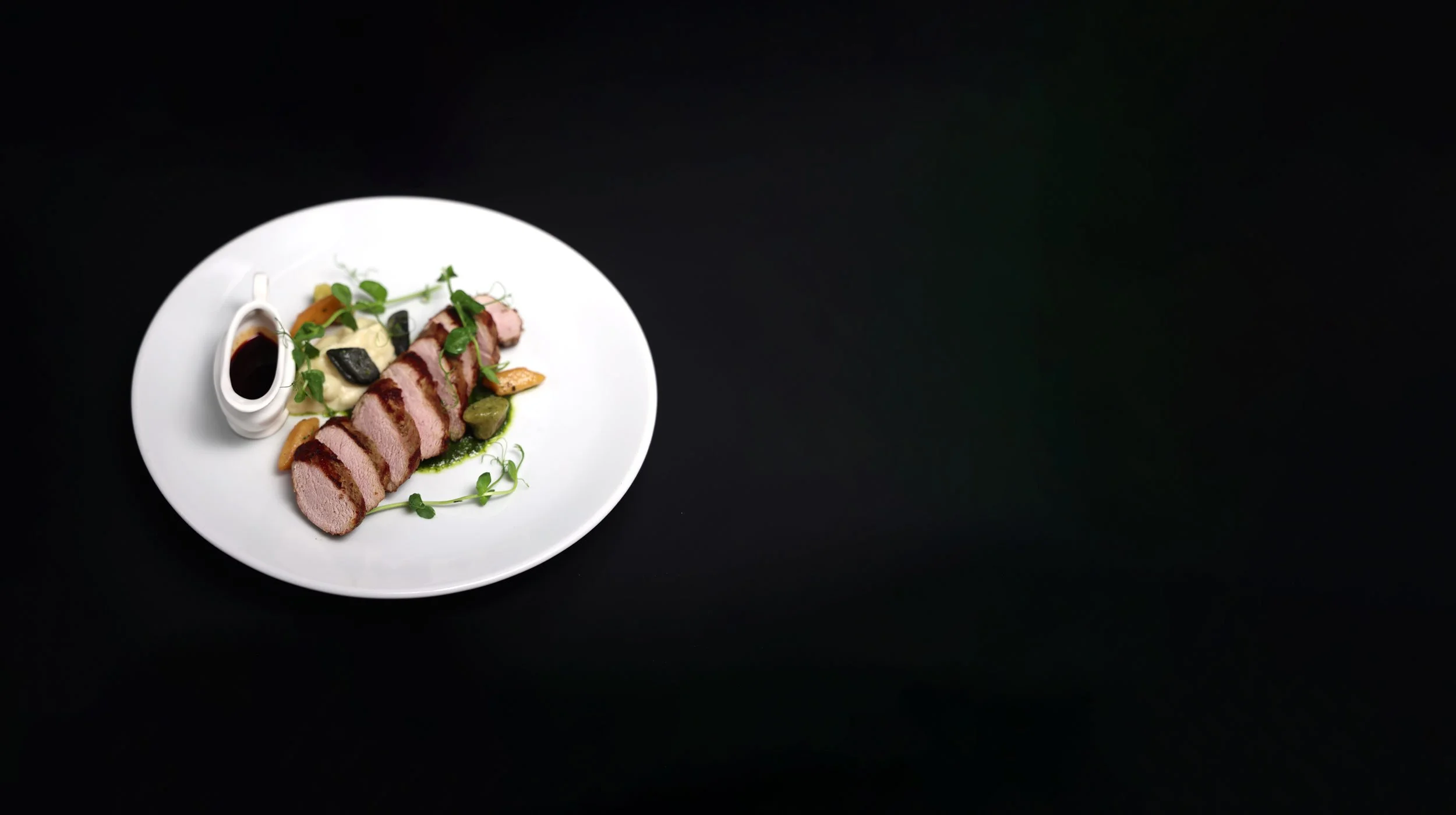 A white plate with slices of cooked meat topped with sauce, garnished with herbs, accompanied by vegetables and a small pitcher of sauce, all on a black surface.