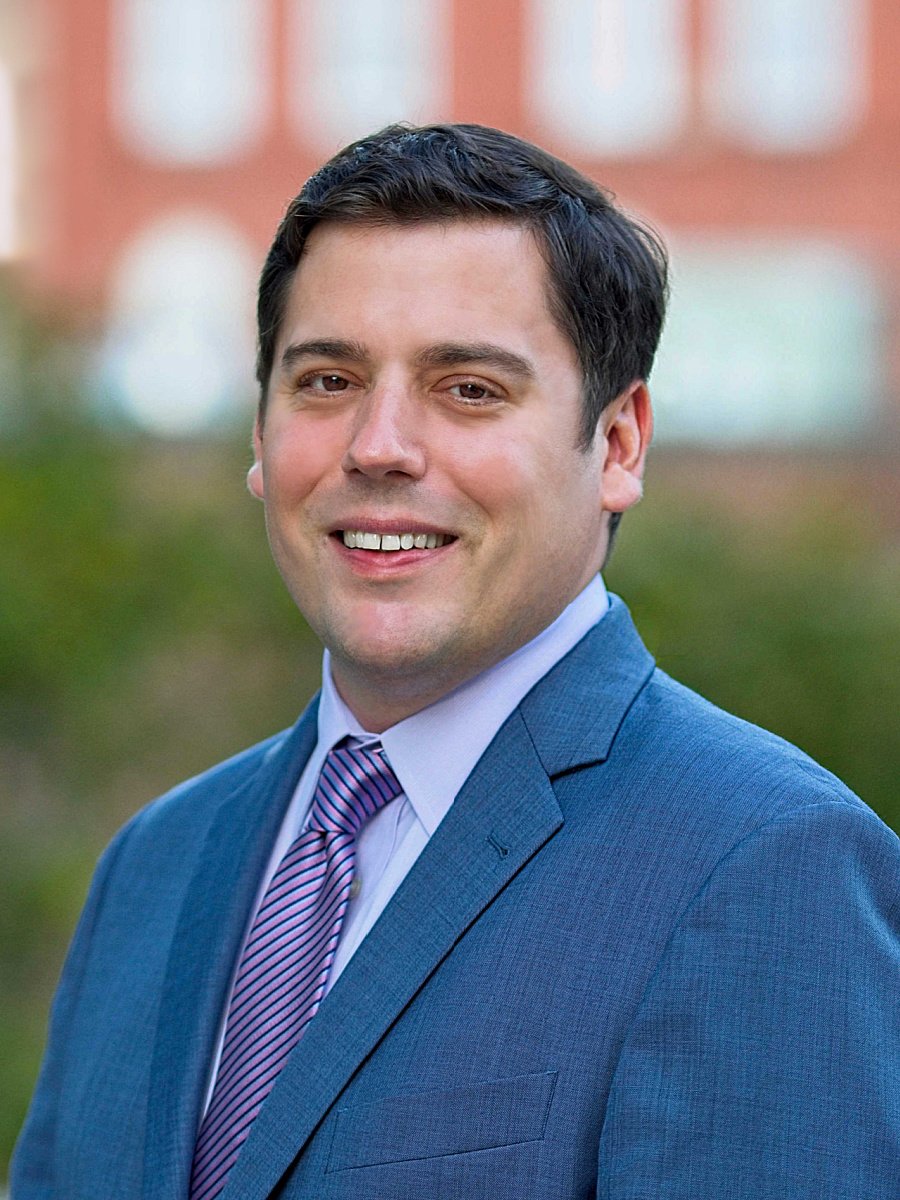 A young man in a blue suit with a white shirt and a pink striped tie, smiling outdoors with a blurred background of trees and a building.