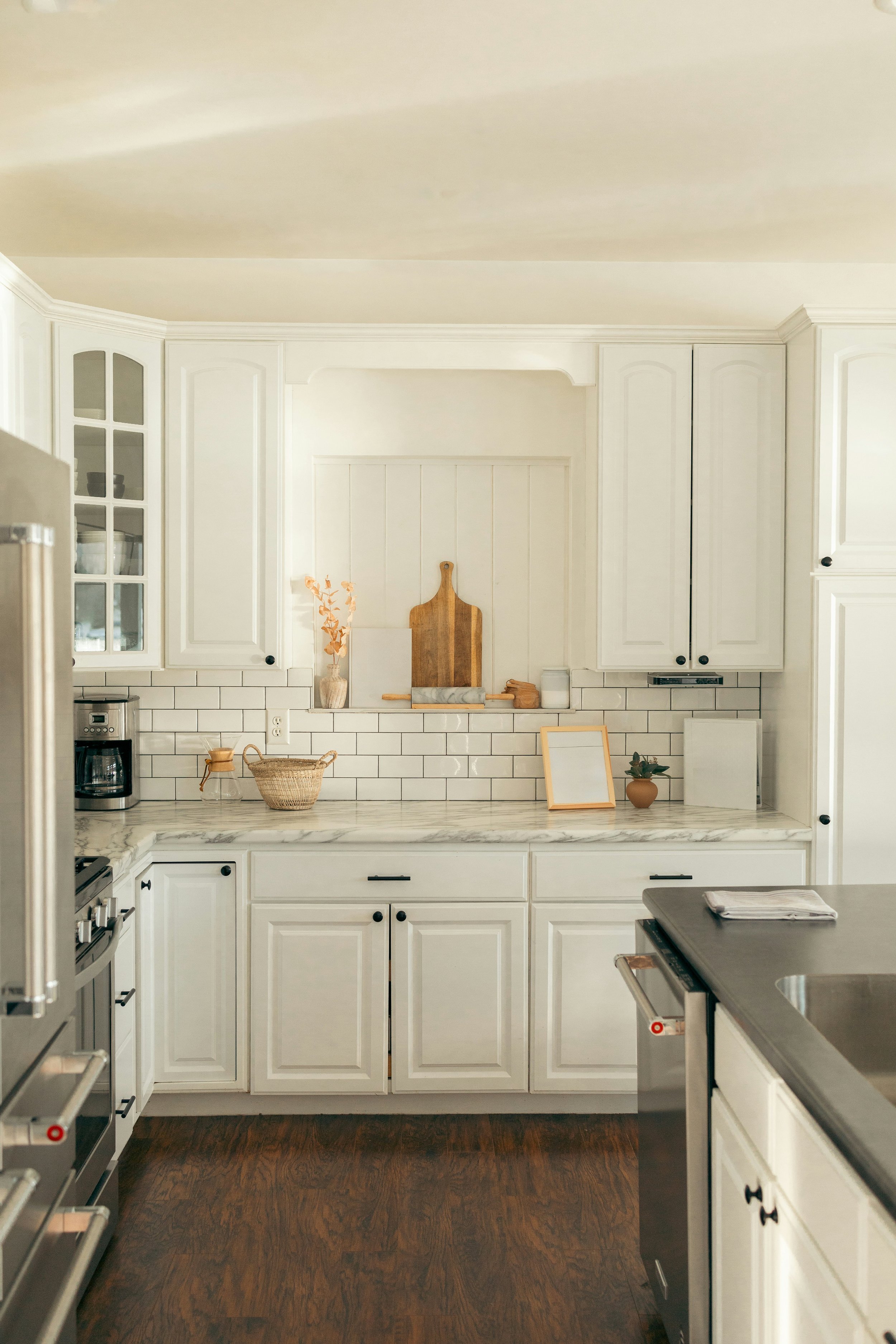 A kitchen with white cabinets, dark wooden floor, marble countertop, white backsplash tiles, and decorative items like a cutting board, vase, and framed print.