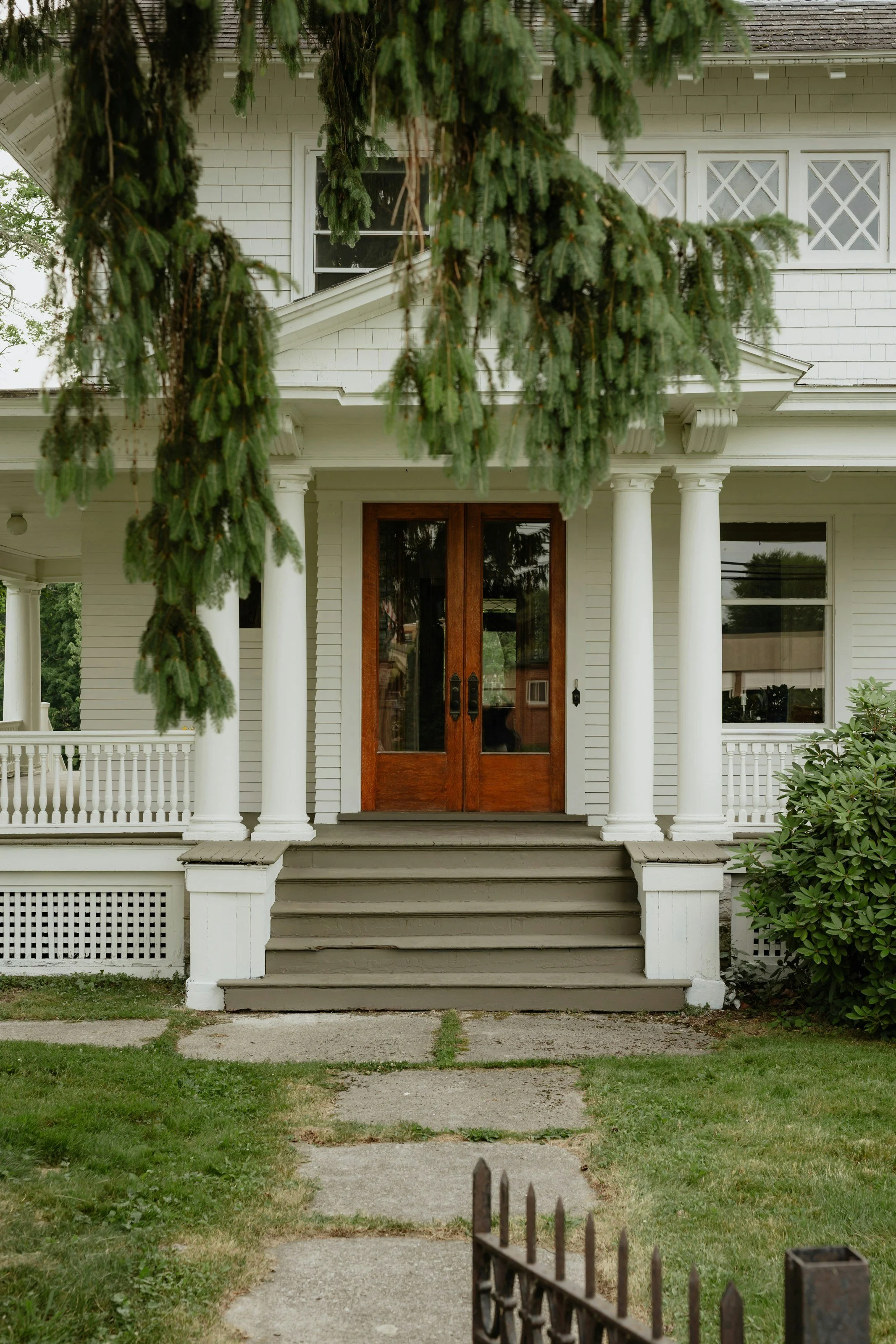 Front porch of a white house with six steps leading up to large wooden double doors, flanked by white columns. A green yard with weeds and a small iron fence in the foreground, with tree branches hanging above.