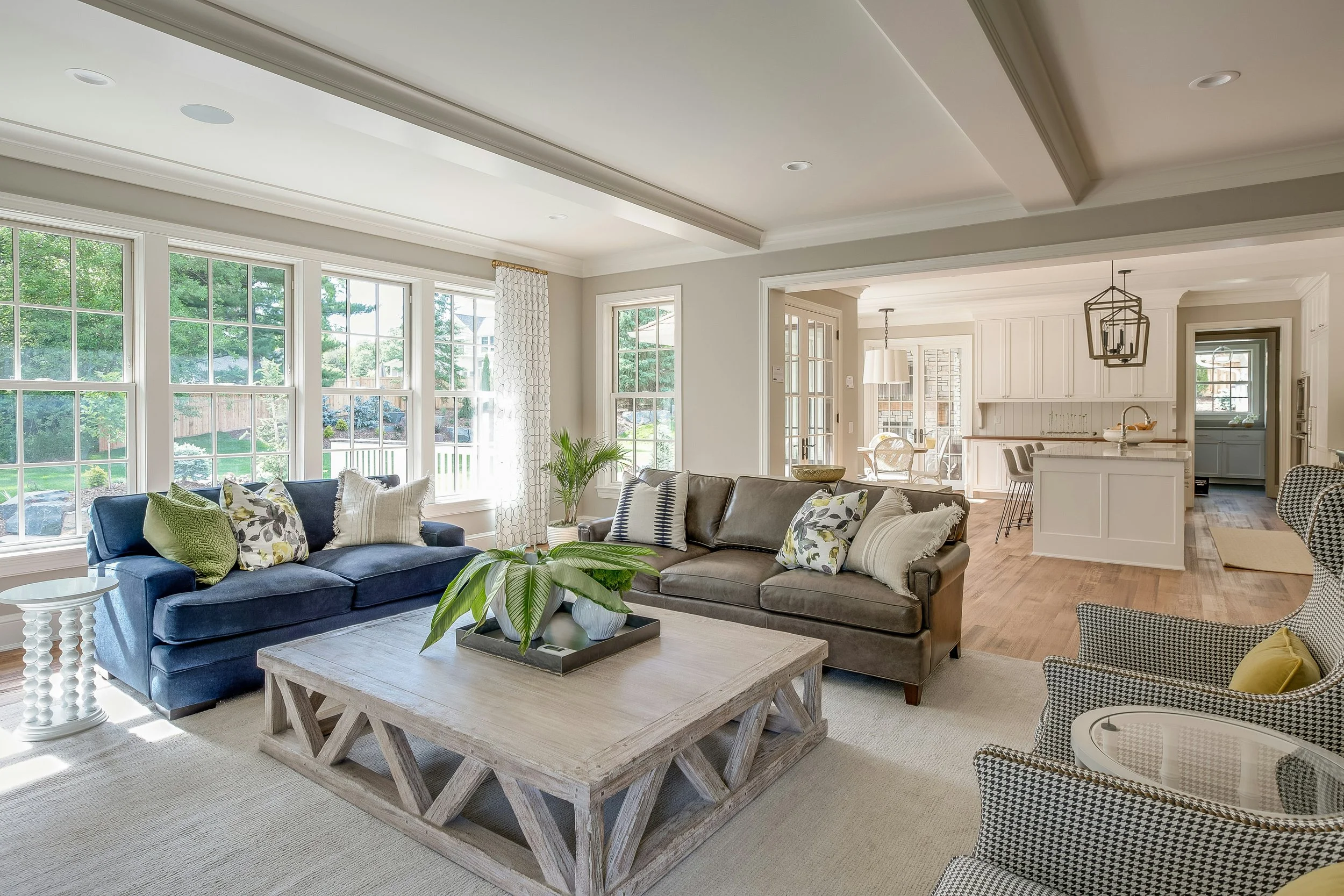 Living room with large windows, white walls, and a mix of blue, gray, and patterned furniture, including a coffee table with a plant, and an open kitchen area in the background.
