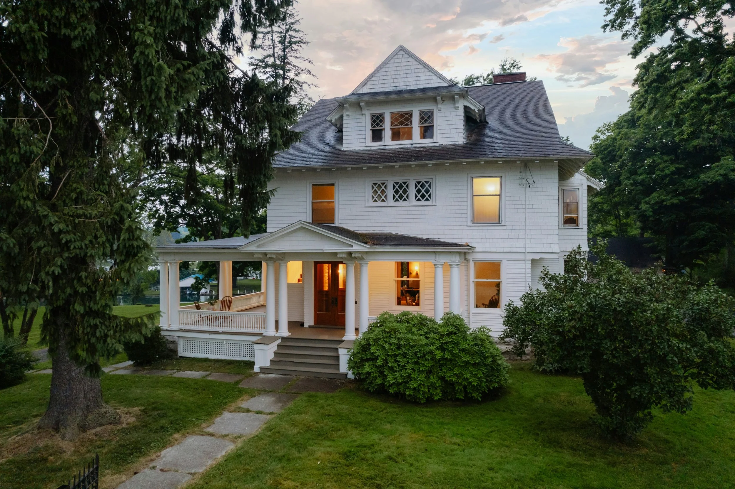 A large white Victorian-style house with a front porch, surrounded by lush greenery and trees, during dusk with lights on inside the house.