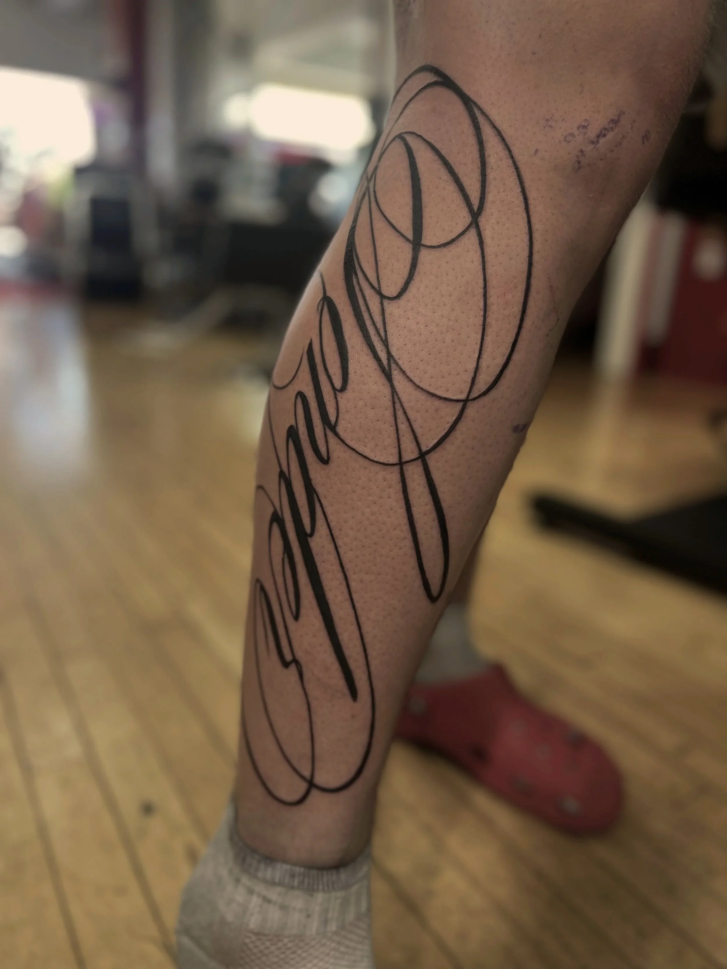 Close-up of a person's arm with a black line art tattoo and small additional text, wearing a gray fishnet sleeve, in an indoor setting with blurred background.