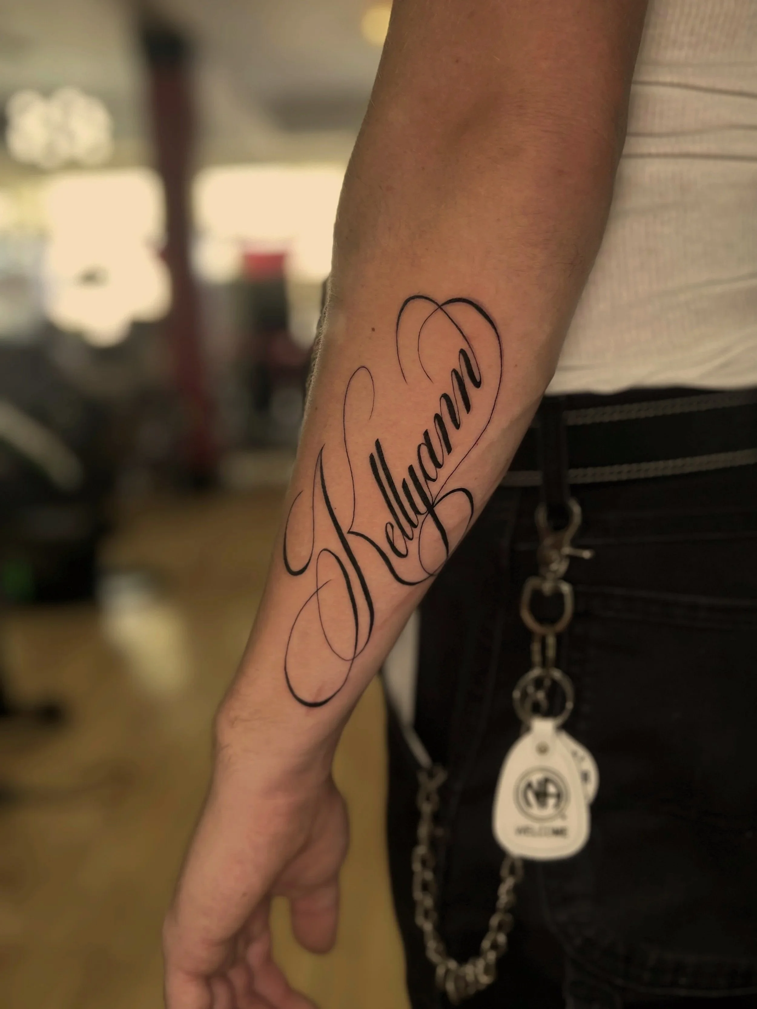 Close-up of a person's arm with a tattoo of the name 'Julianne' inside a heart outline, with a blurred background and part of a black purse with silver tags hanging from it.