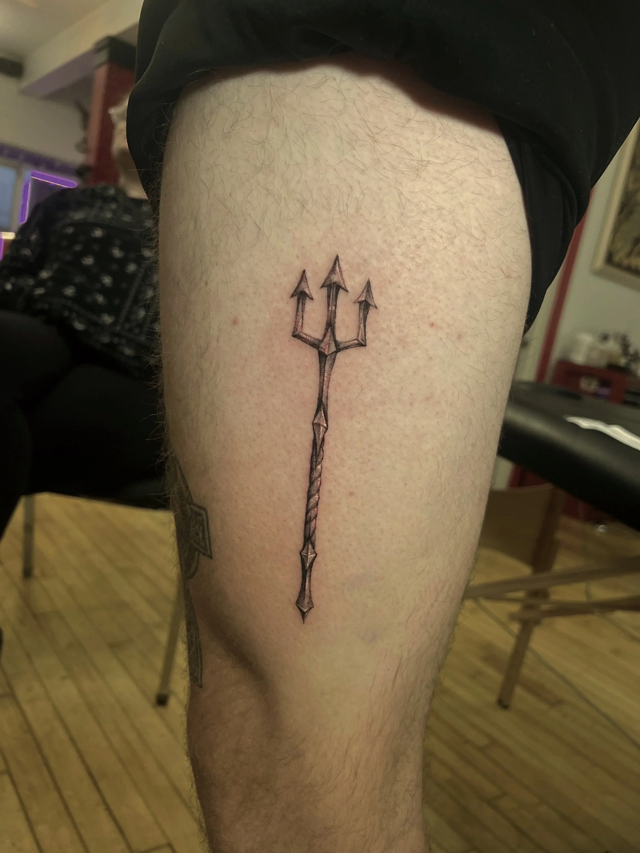 Tattoo of a black outlined trident on a person's thigh.