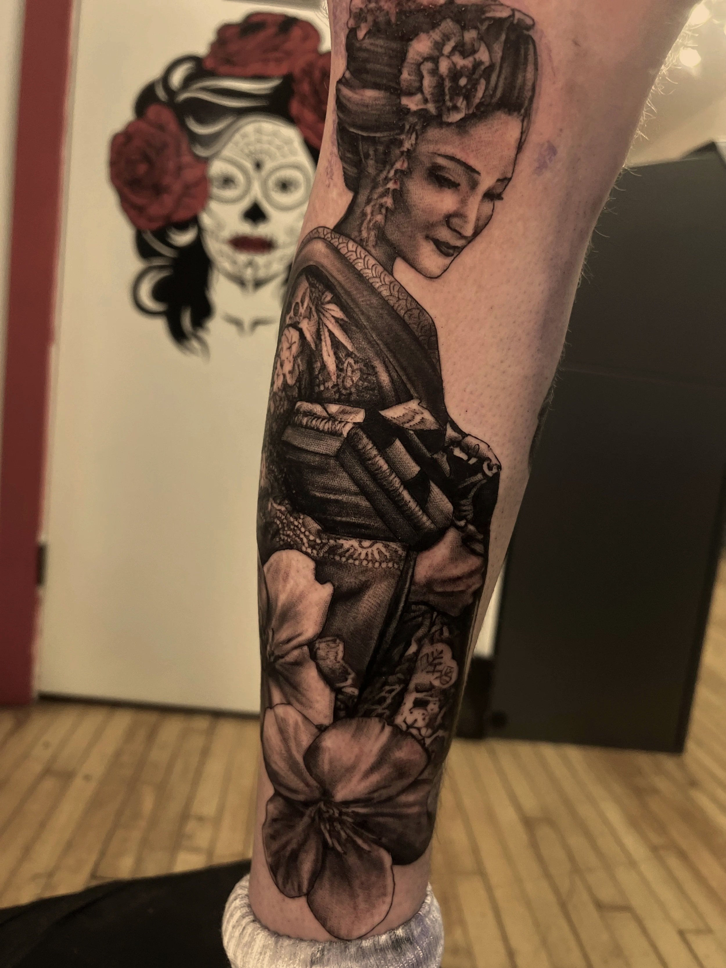 A monochrome tattoo of a woman with styled hair, holding a handgun, on someone’s arm, with a background of a wall with a decorative mask and flowers.