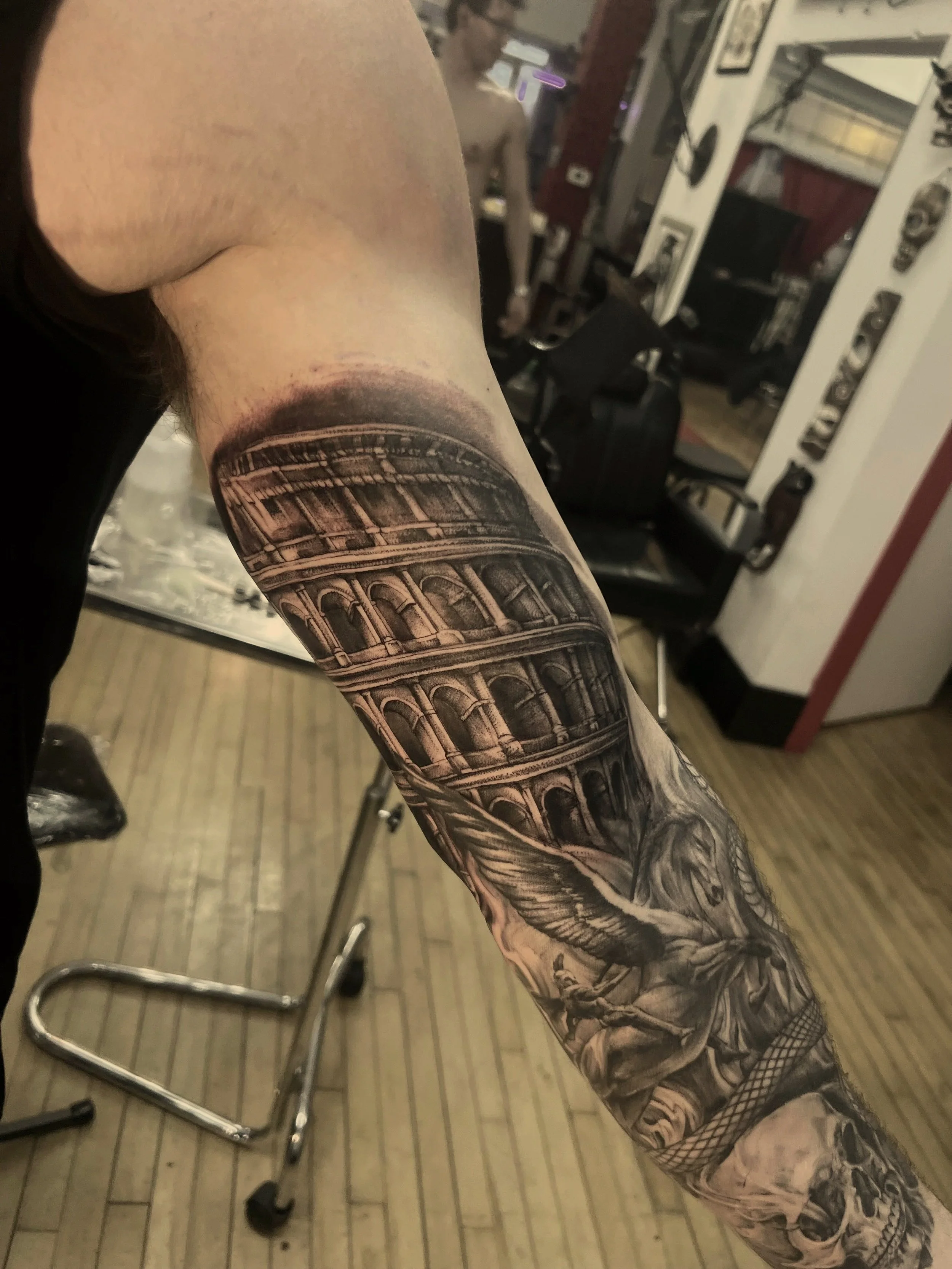 Tattooed arm with architectural design resembling the Colosseum and a skull, in a tattoo parlor with tattoo equipment and a person in the background.