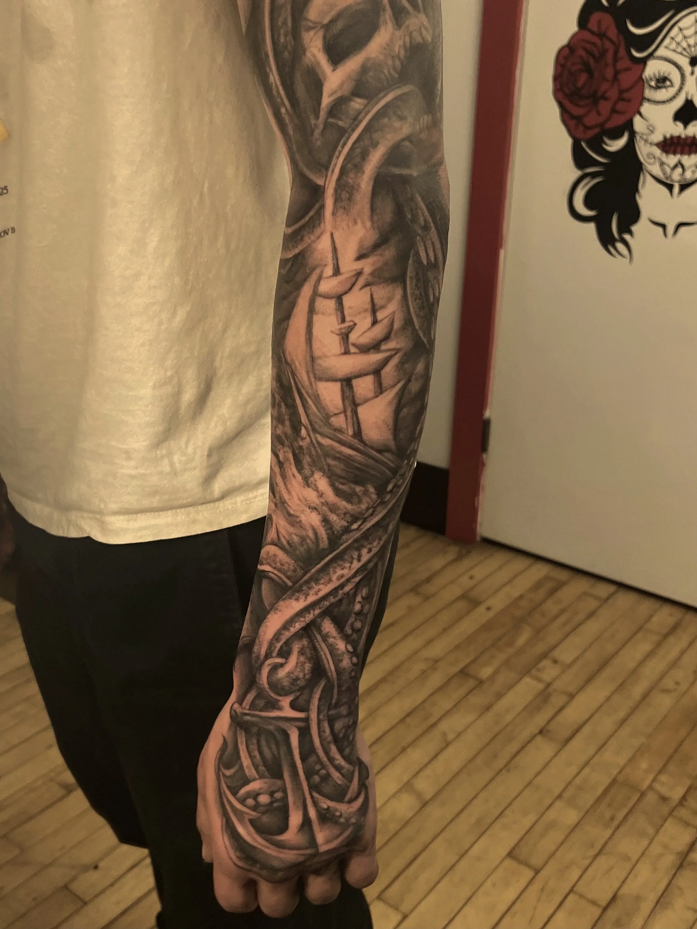 Close-up of a person's arm and hand, showing an intricate black and gray tattoo sleeve with a snake and various decorative patterns. The person is wearing a short-sleeved, light-colored shirt.