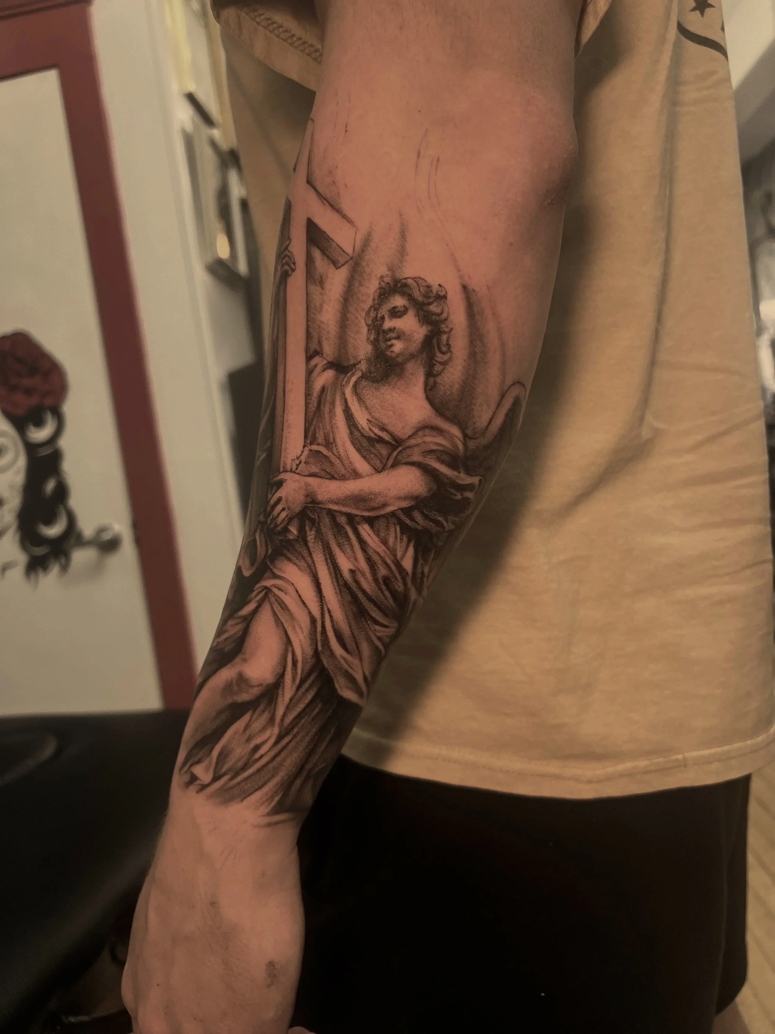 Tattooed forearm and hand of a person showing a woman with curly hair and a smile, holding a large object, with a cross in the background.