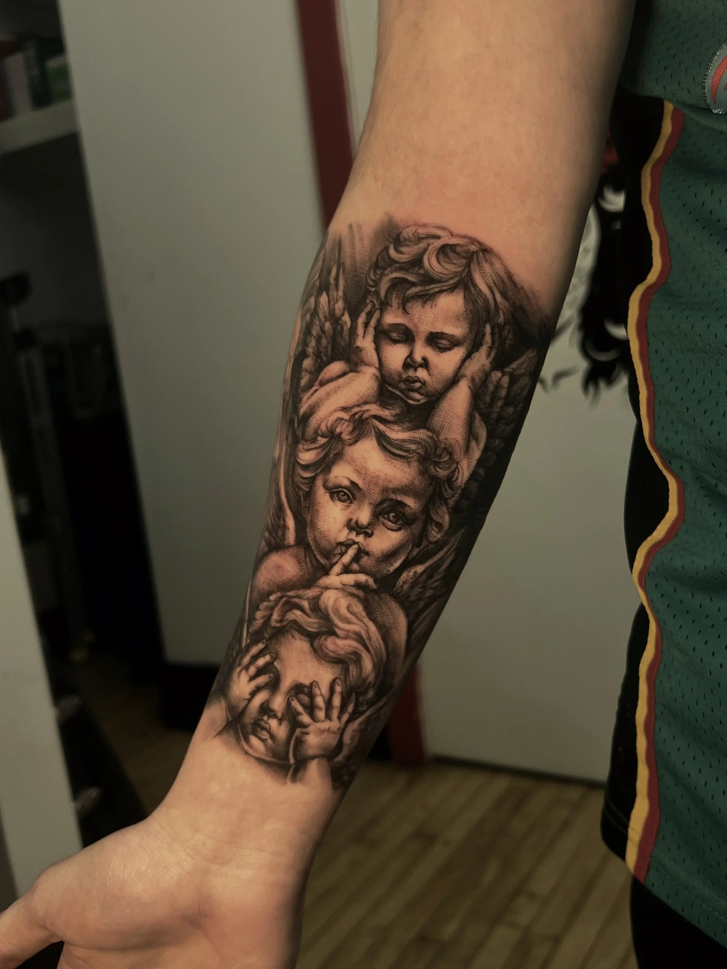 A detailed black and gray tattoo of cherubs or angels on a person's forearm, featuring two faces of cherubs with wings and hands covering their ears.